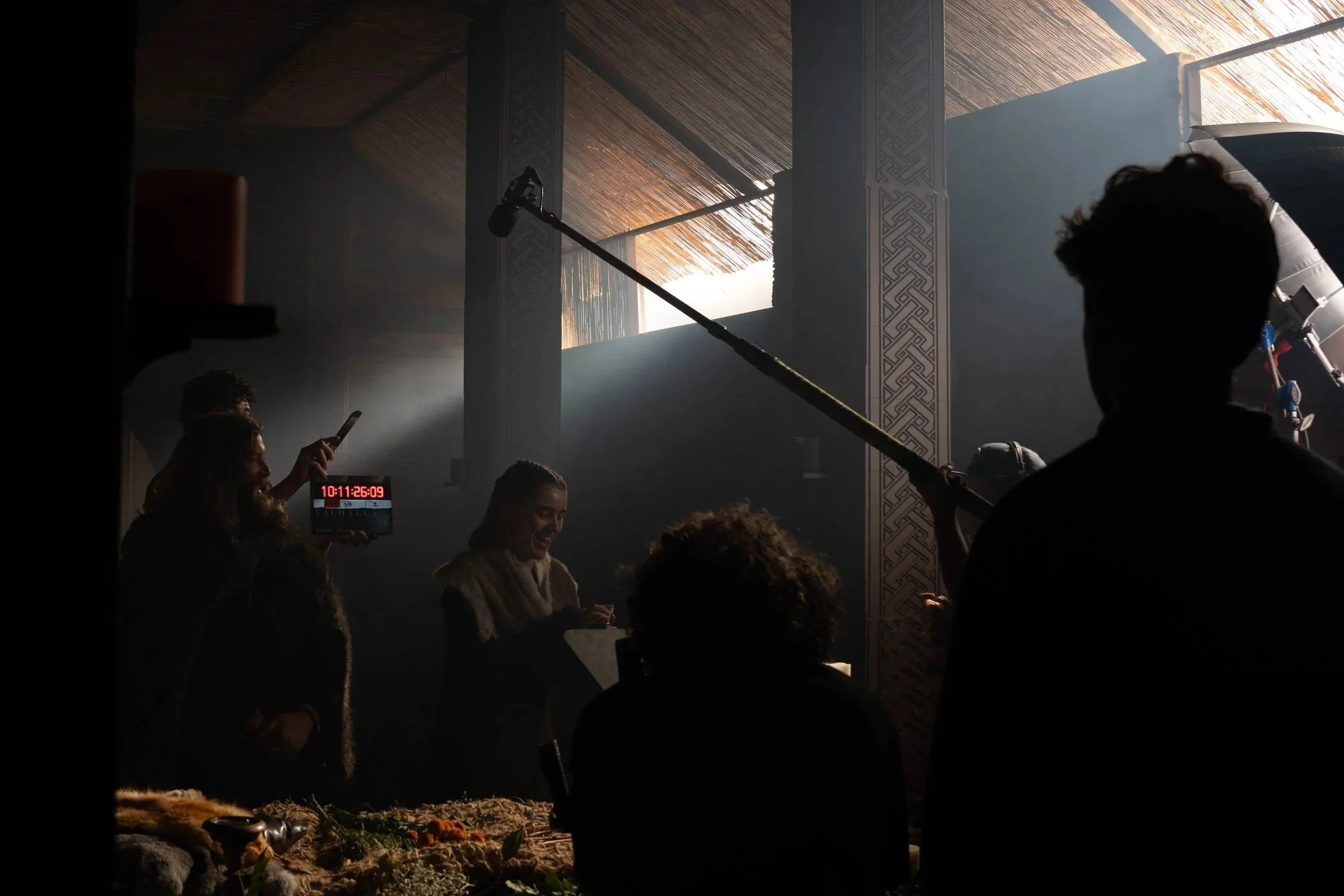 Scene of a production set or event with people, equipment, and decorative pillars, illuminated by light through a window or opening in the ceiling.