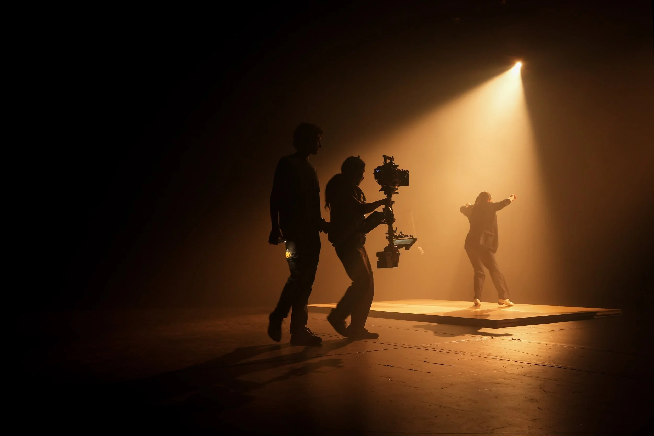 People on stage with dramatic lighting, one person filming with a professional camera, and others performing or posing.