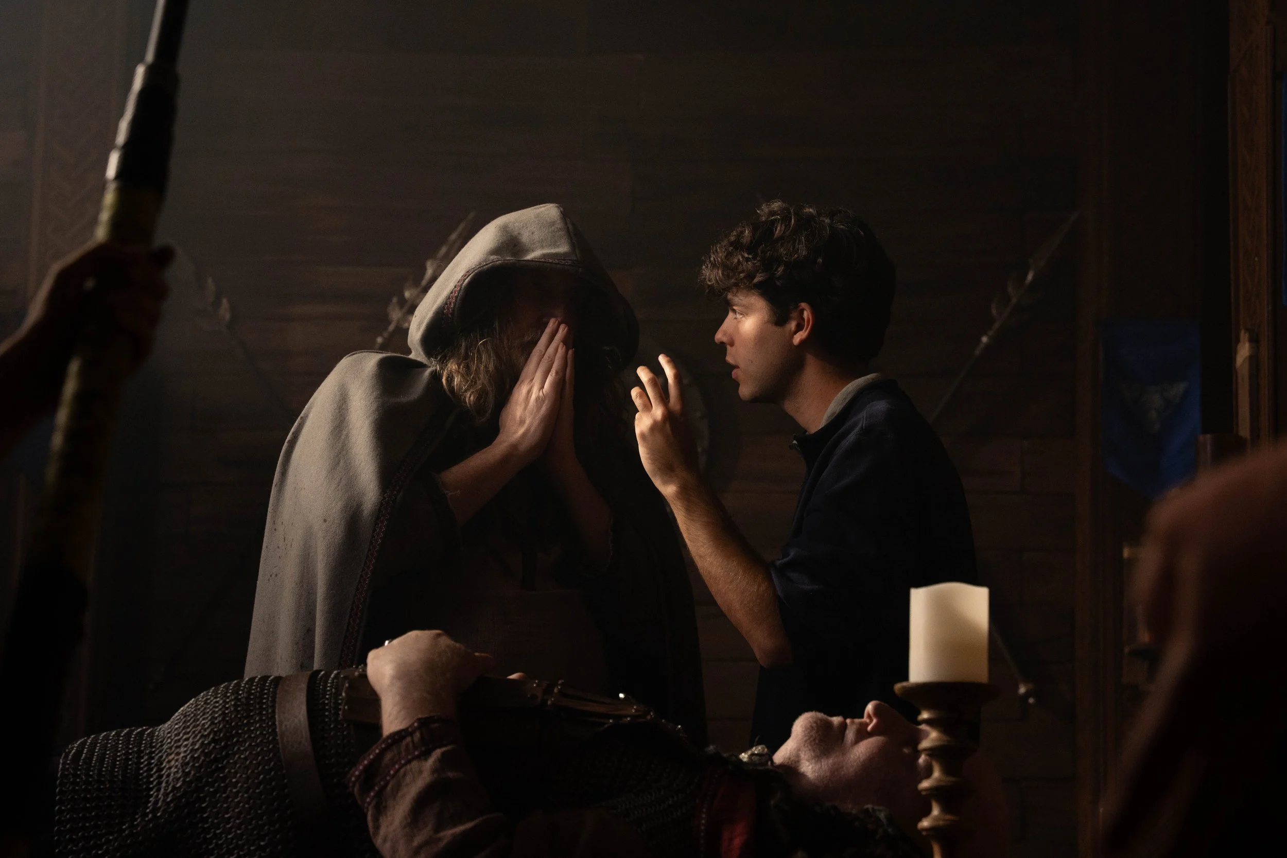 A young man and woman are sitting close together in a dimly lit room, engaging in a private conversation. The woman is wearing a hoodie and has her hands pressed against her face, while the man is leaning in, speaking softly. In the background, a person is holding a paddle or stick, and there is a lit candle on the table in front of them.