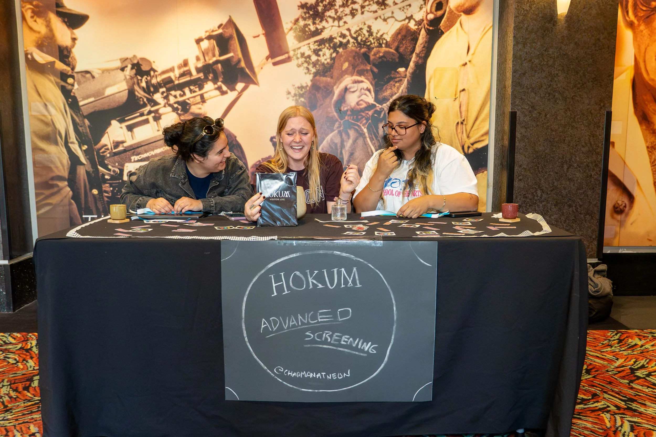 Three women sitting at a table with a black tablecloth with a chalkboard sign reading 'HOKUM DANCER ADVANCED SCREENING' in front. One woman holds a bag with 'HOKUM' written on it and is laughing, the other two are engaged and looking at her. A large, sepia-toned nostalgic background photo depicting film production equipment and older people is behind them.