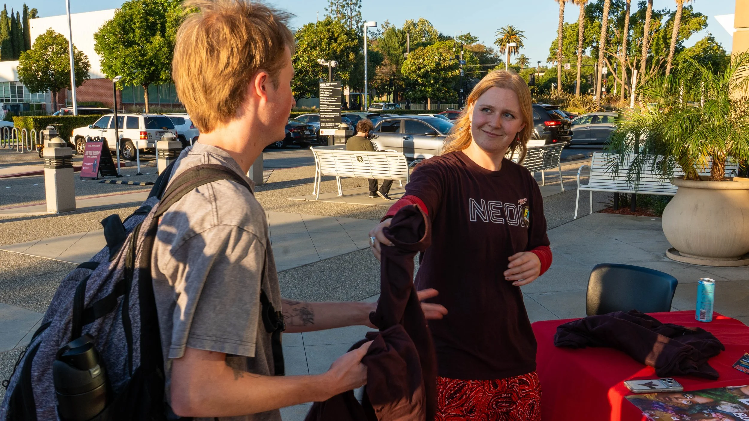Two young women are conversing outdoors next to a table with various items, with a parking lot and trees in the background. One woman has short blonde hair and a backpack, while the other has long red hair and is wearing a black sweatshirt.