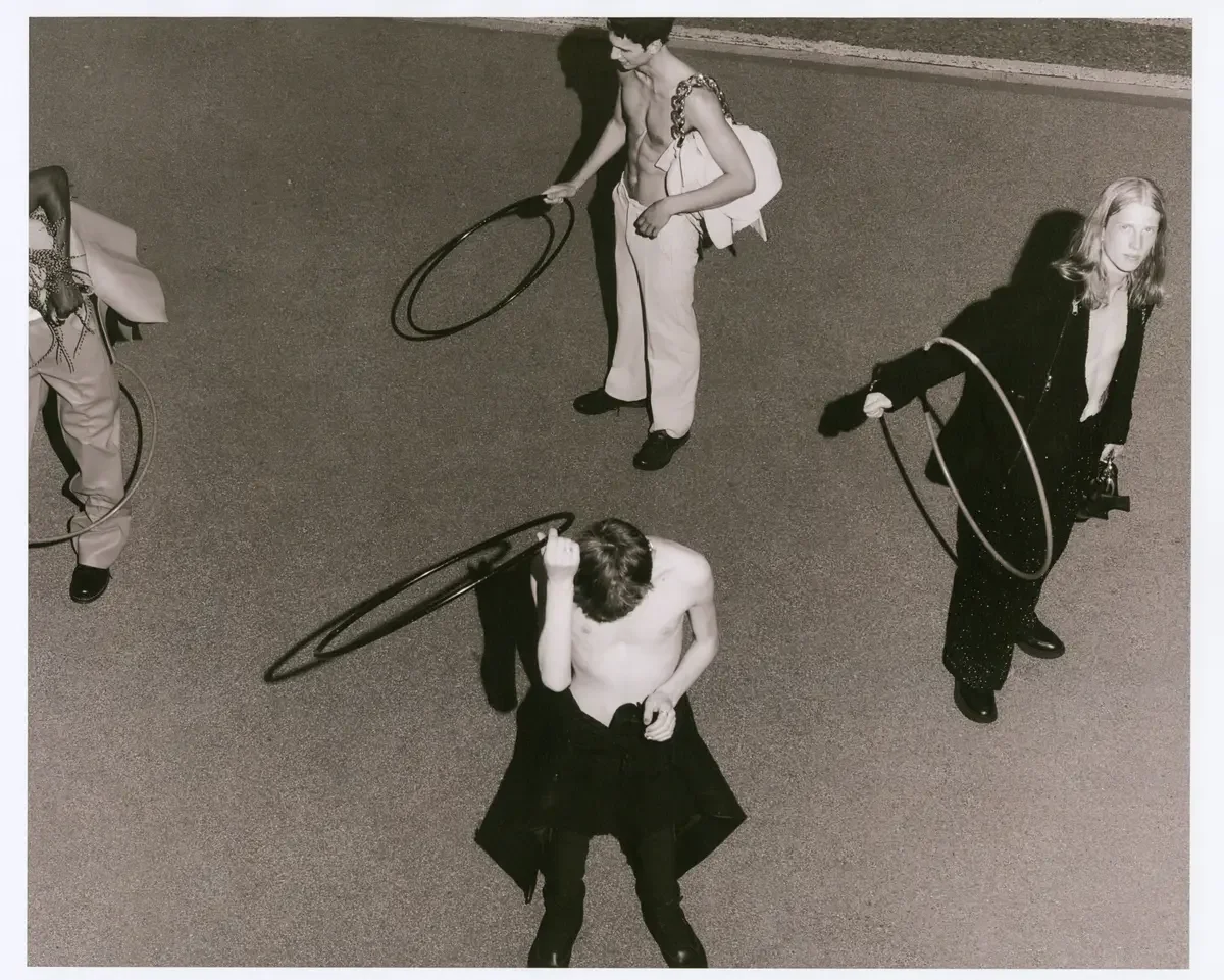 A black and white photo of four young people standing on the street, each holding a hula hoop. One person is shirtless, another wears a blazer, and others are dressed casually. The photo is taken from above.