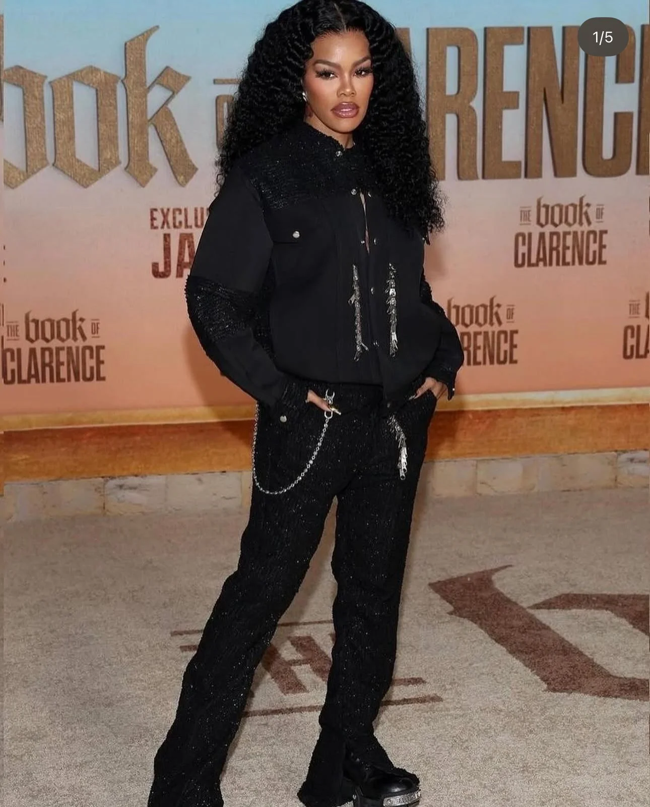 A woman with long, curly black hair, wearing black sparkly pants, a matching black jacket with sparkly accents, black shoes, and accessorized with chains and rings, posing in front of a promotional backdrop for 'The Book of Clarence'.