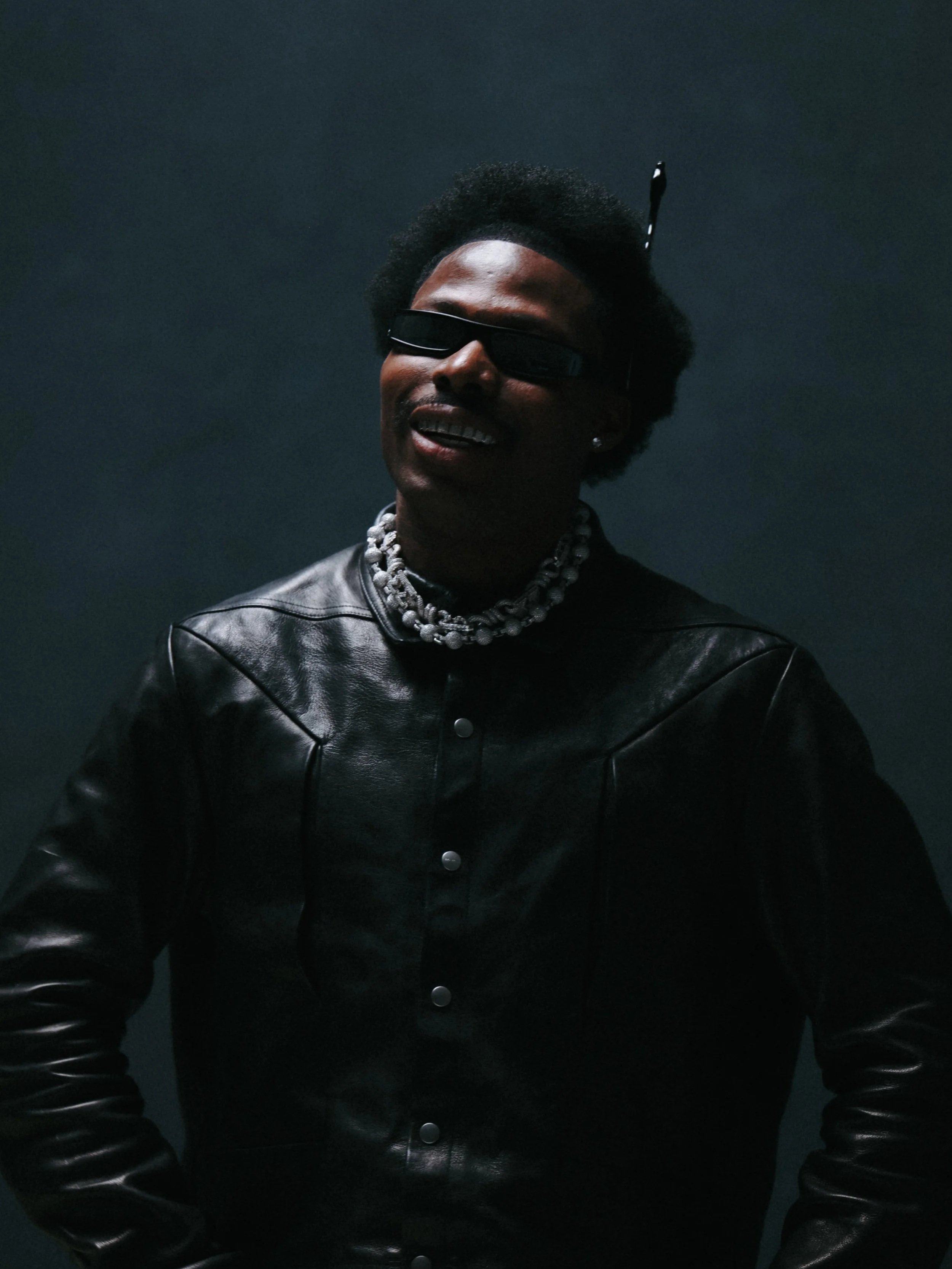 Person wearing sunglasses, black leather jacket, and chunky jewelry, smiling against a dark background.