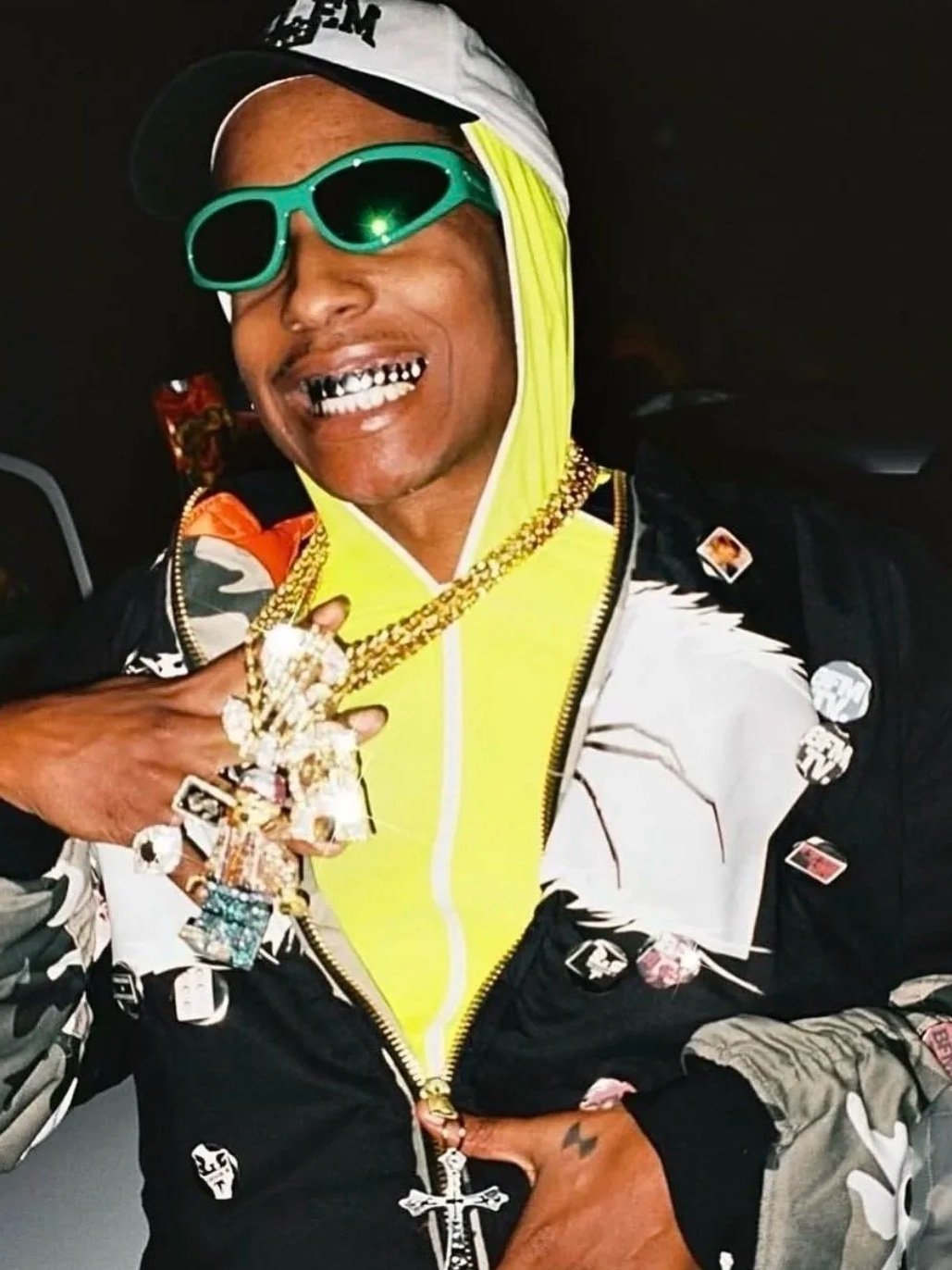 Person wearing a yellow hoodie, black and white jacket, green sunglasses, and a cap. They have gold teeth, multiple rings, and large jewelry, including a chain with a cross. The person is smiling and appears to be in a dark setting.