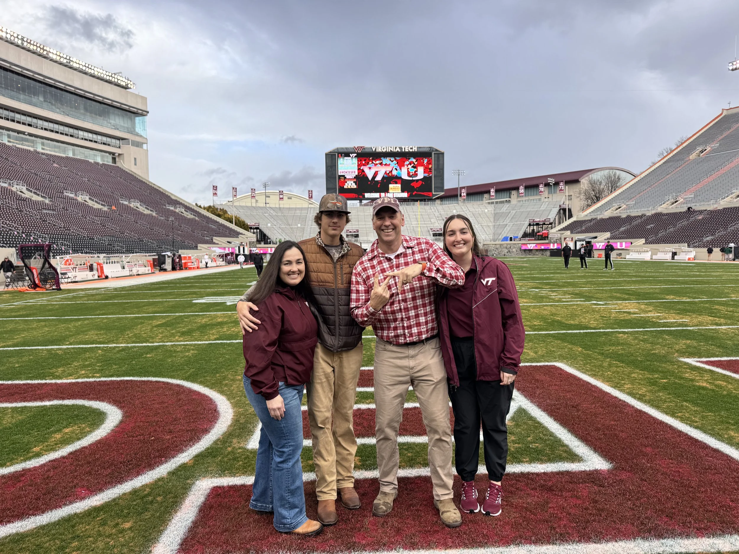 Four people standing on a football field at Virginia Tech stadium with a large digital scoreboard behind them showing 'Virginia Tech' and 'Welcome to Lane Stadium' during daytime.