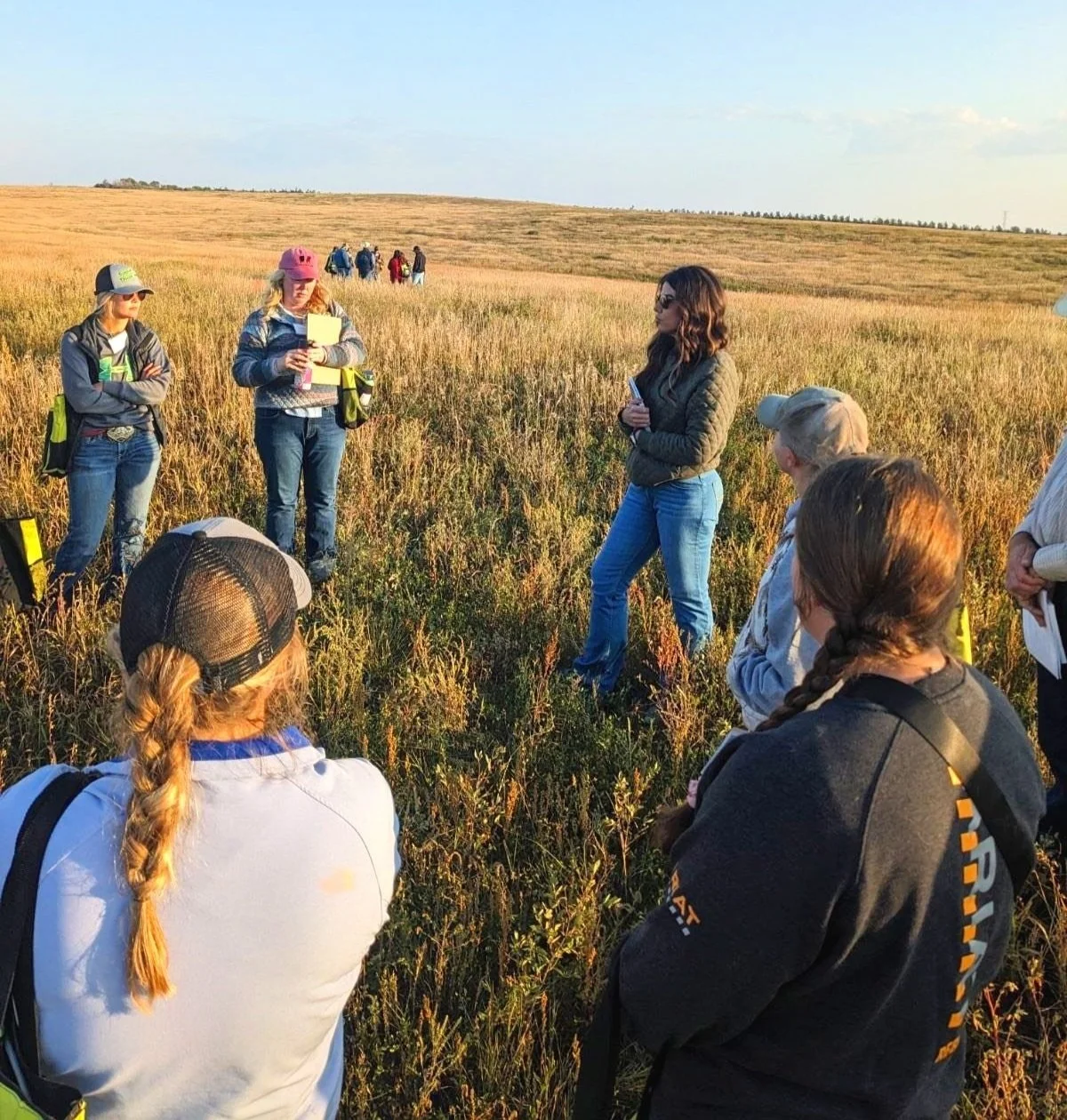 A group of people standing in a field, having a discussion. Some are wearing hats and holding clipboards. The setting appears to be outdoor, with tall grasses and a clear sky in the background.