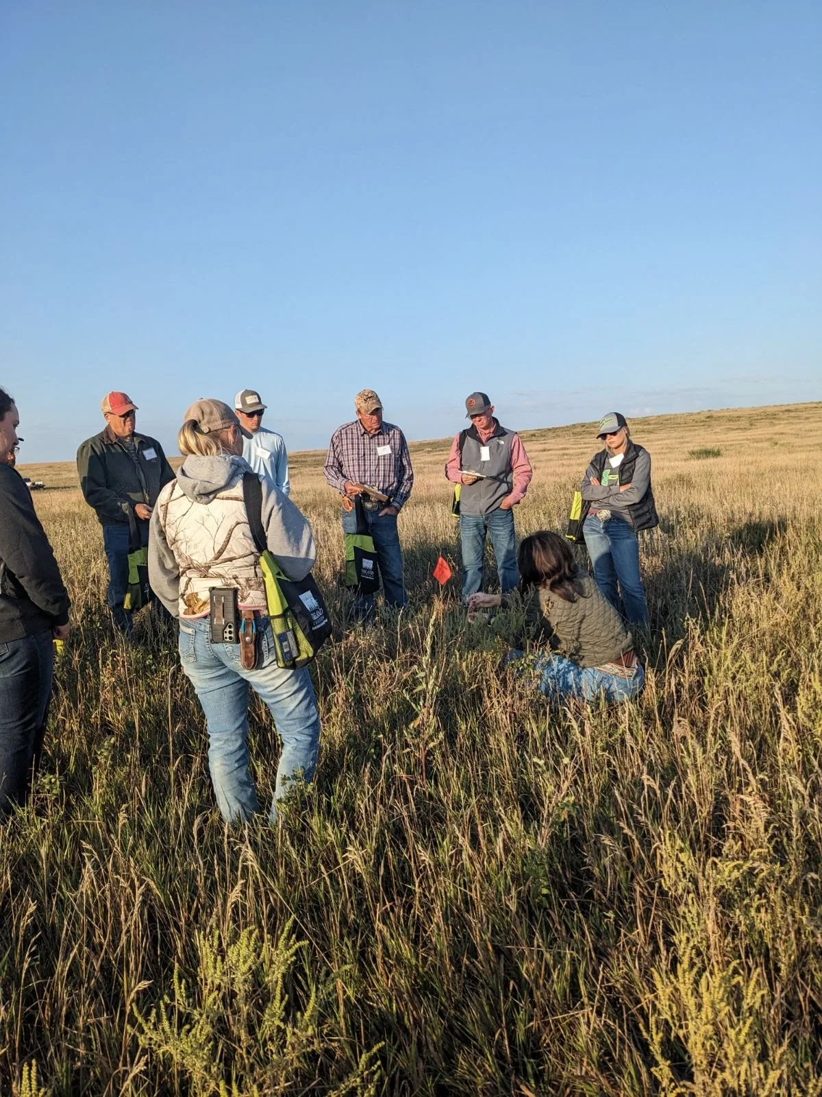 A group of people standing in a grassy field, participating in an outdoor activity or workshop, with some holding notebooks and wearing casual clothing.