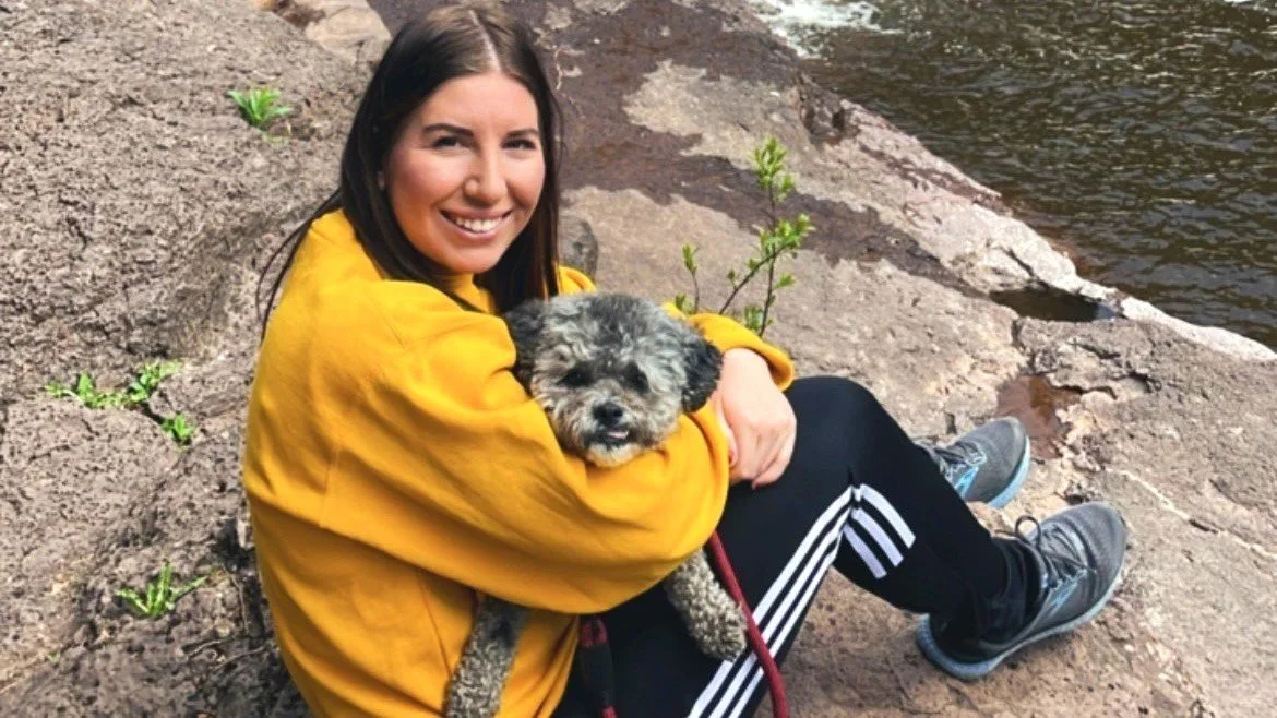 Lady wearing a yellow jumper sitting by a lake on rocks holding a Shih-Poo dog