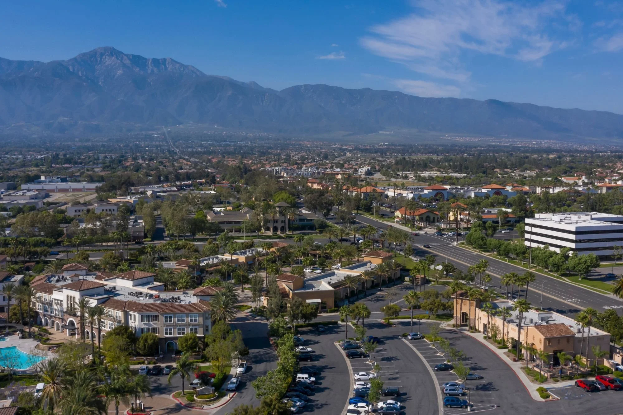 Aerial view of a suburban area with buildings, palm trees, streets, and parking lots, with mountains in the background under a partly cloudy sky.