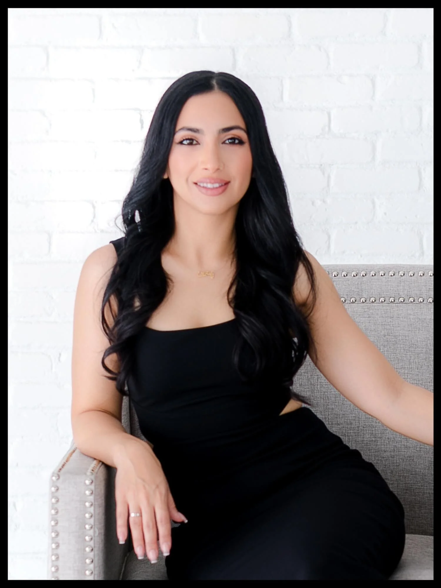 A woman with long black hair, wearing a black sleeveless dress, sitting on a beige armchair with silver nailhead trim against a white brick wall, smiling at the camera.