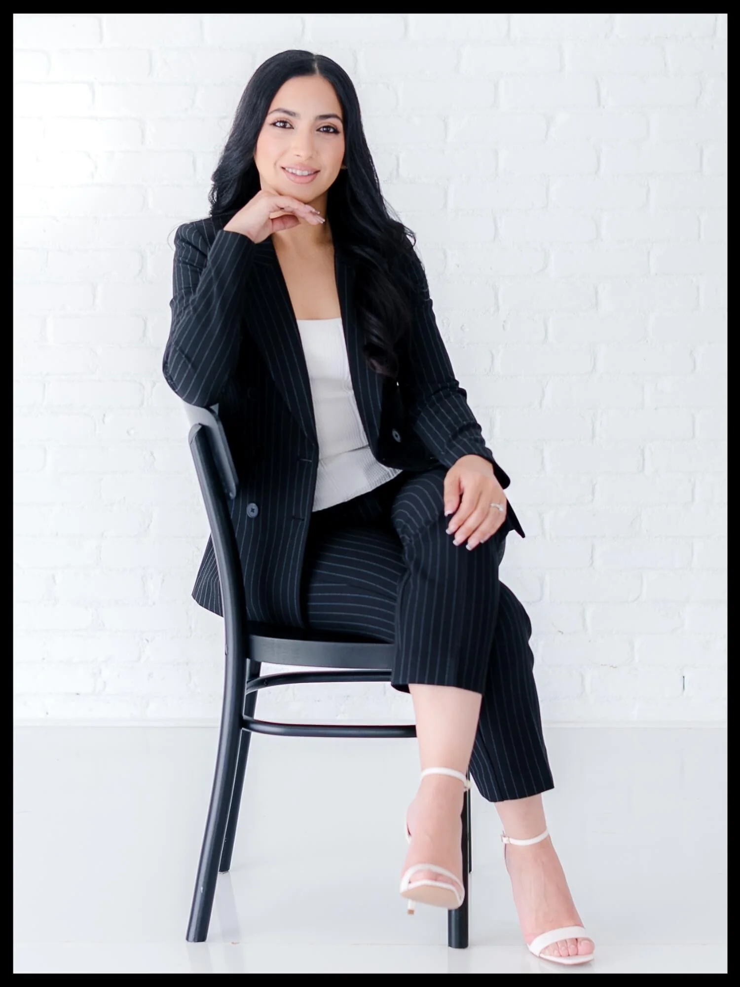 A woman with long black hair, wearing a black pinstripe suit and white top, sitting on a black chair against a white brick wall, smiling, with one hand resting on her face and the other on her knee. She is wearing white high heel shoes.