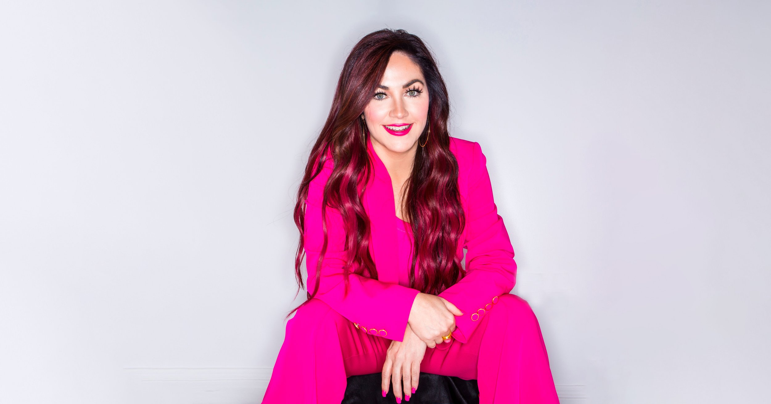 Woman with long, wavy, dark red hair wearing a bright pink suit, sitting with one knee up and smiling at the camera, against a plain white background.