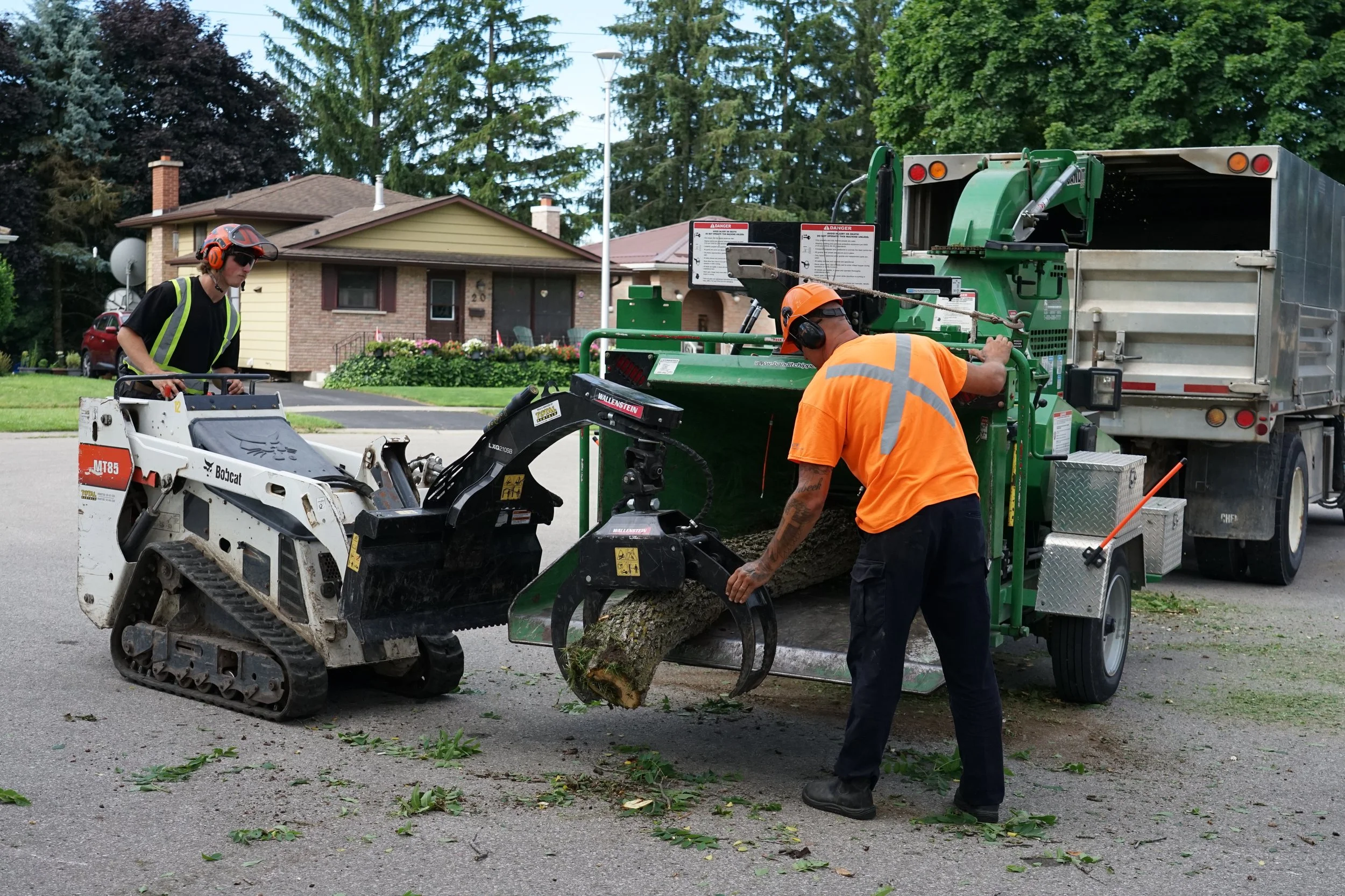 Two workers in safety gear cut a tree log with a wood chipper on a residential street.