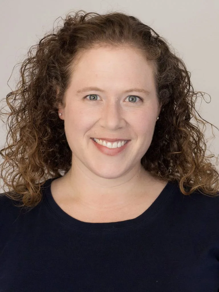Smiling woman with curly red hair and blue eyes wearing a black shirt.