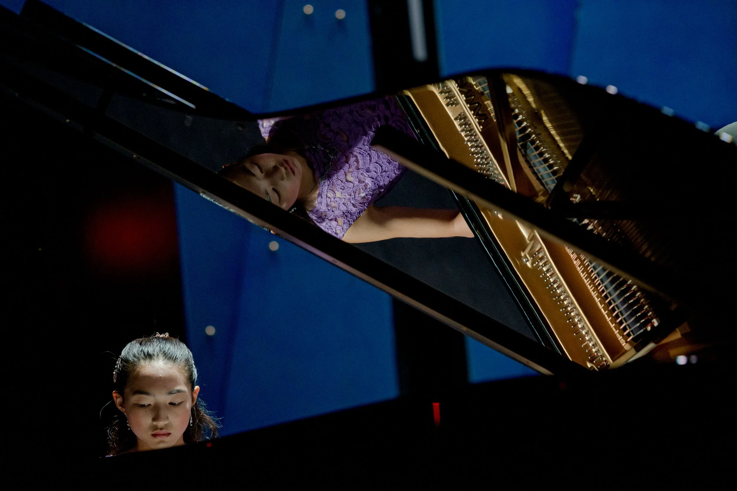A young woman playing a grand piano in a concert setting with blue walls.