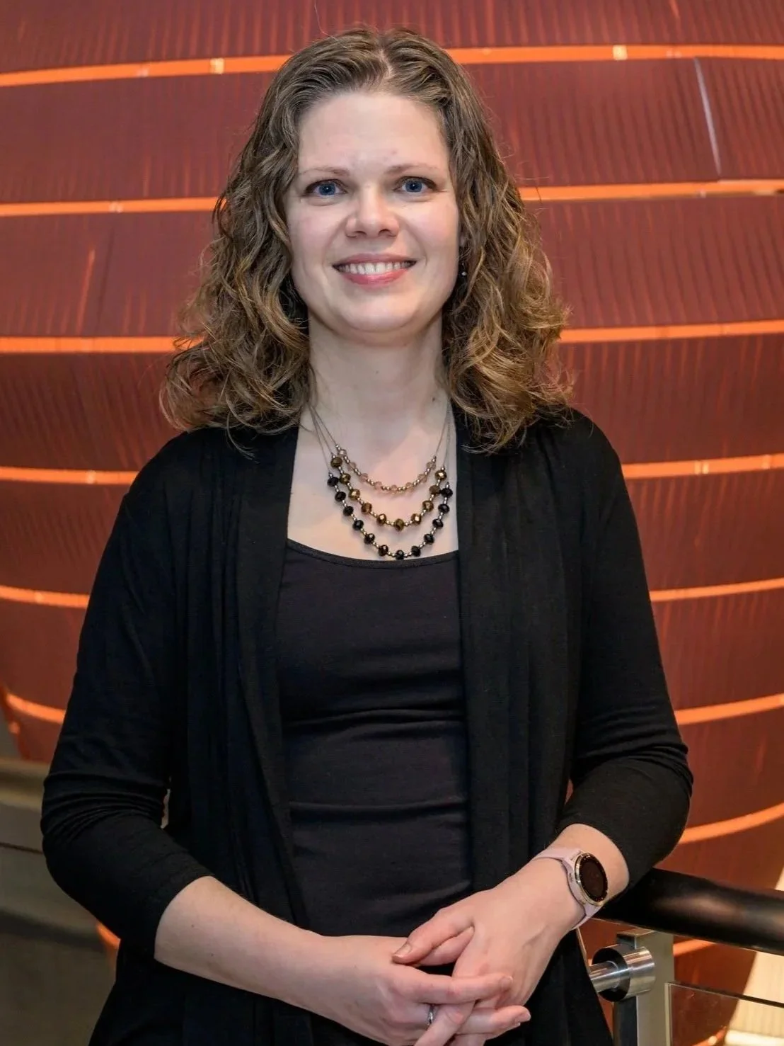 A woman with shoulder-length curly blonde hair and blue eyes standing indoors in front of a wooden wall with horizontal slats. She is wearing a black top, a black cardigan, a multi-strand pearl necklace, and a wristwatch. She is smiling and resting one hand on a railing.