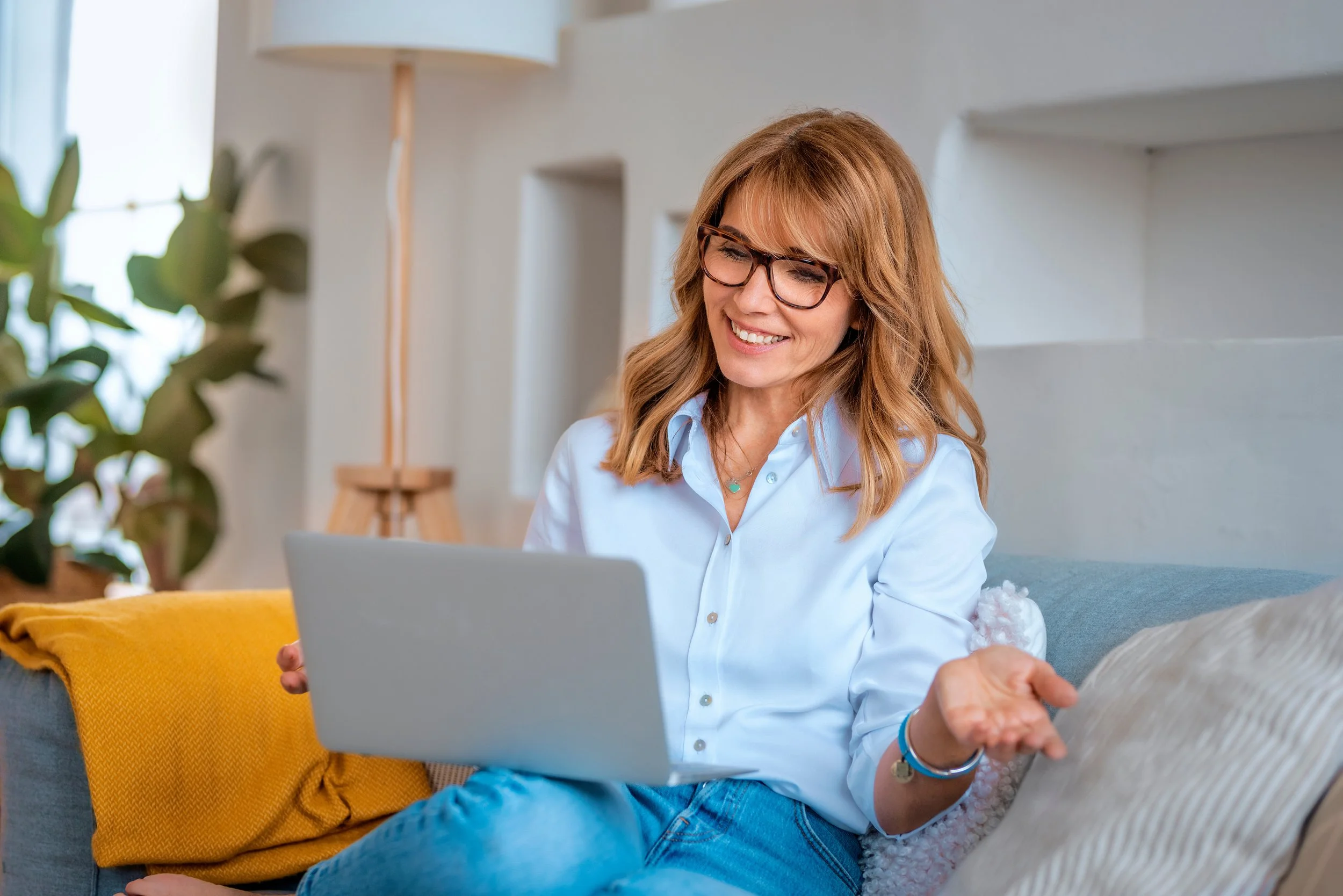 A woman with red hair and glasses sitting on a couch, smiling and looking at a laptop.
