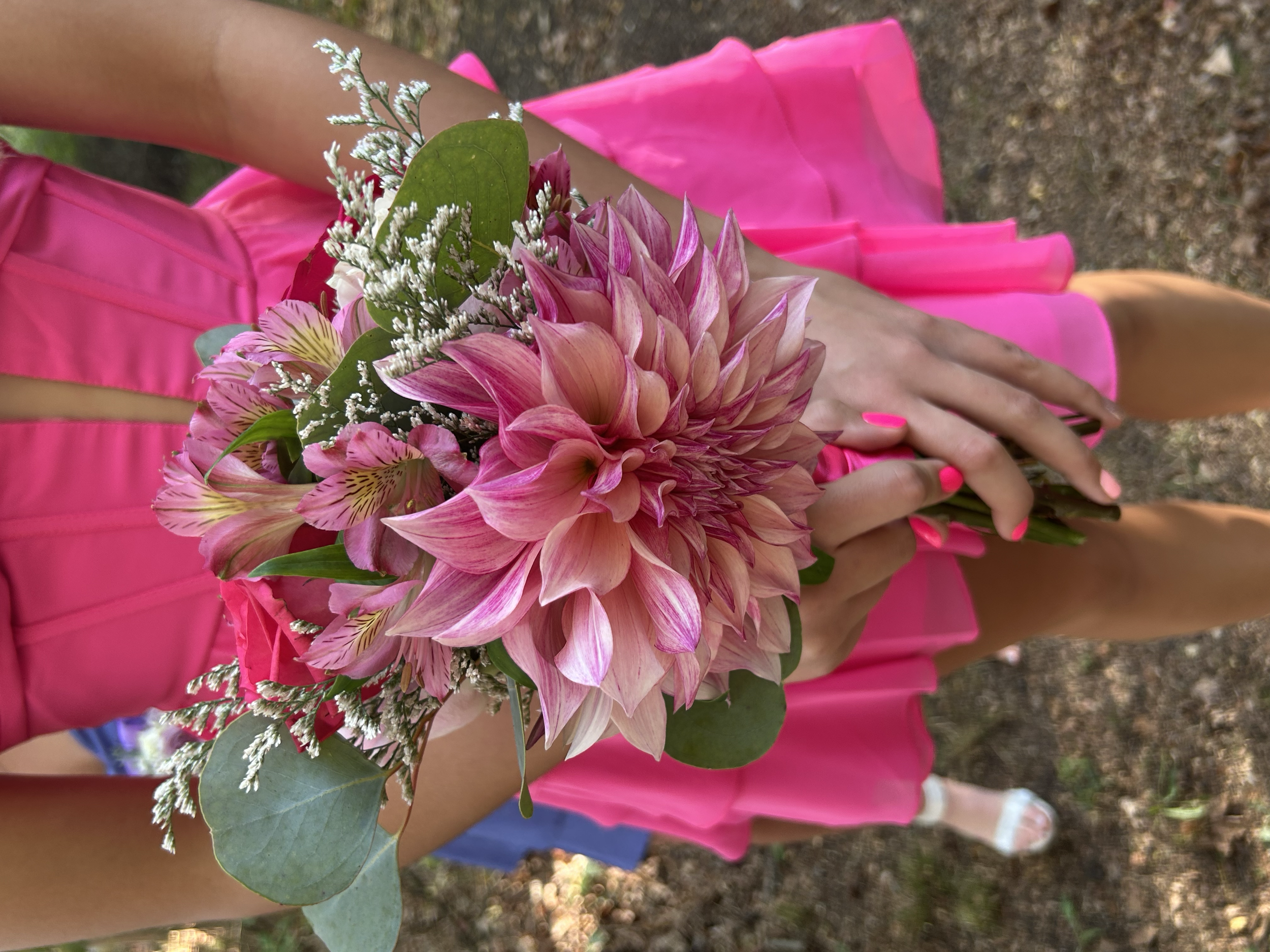A woman wearing a bright pink dress holding a bouquet of pink flowers, including a large dahlia, with greenery and smaller flowers, outdoors on a dirt path.
