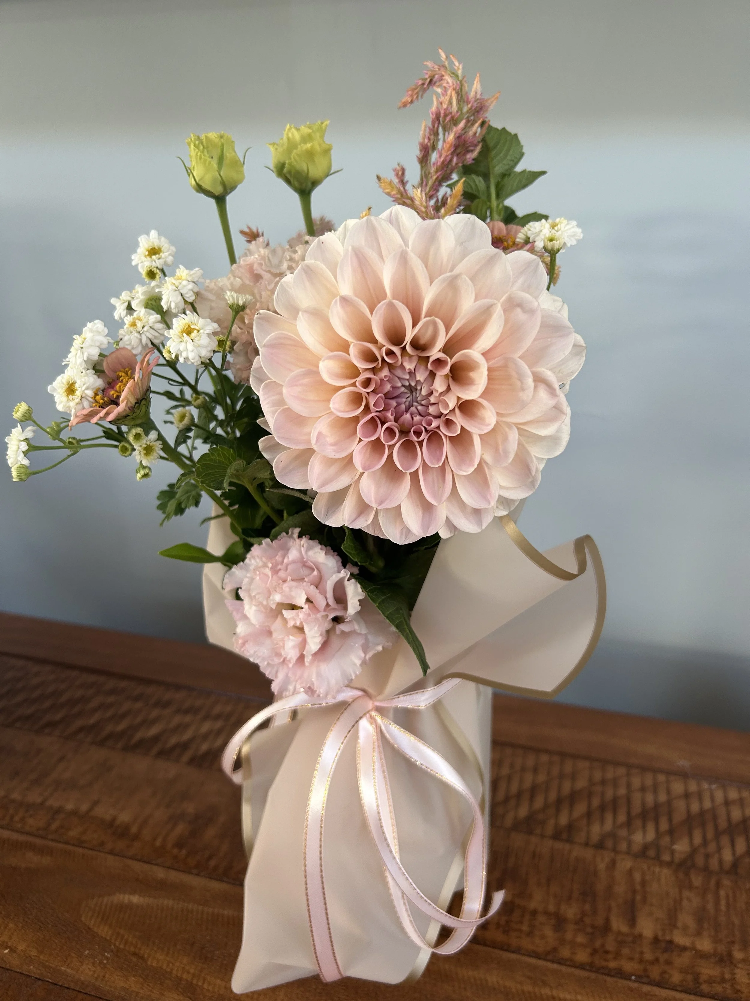 A pink and white flower bouquet in a white paper wrapping, tied with a pink ribbon, placed on a wooden surface.