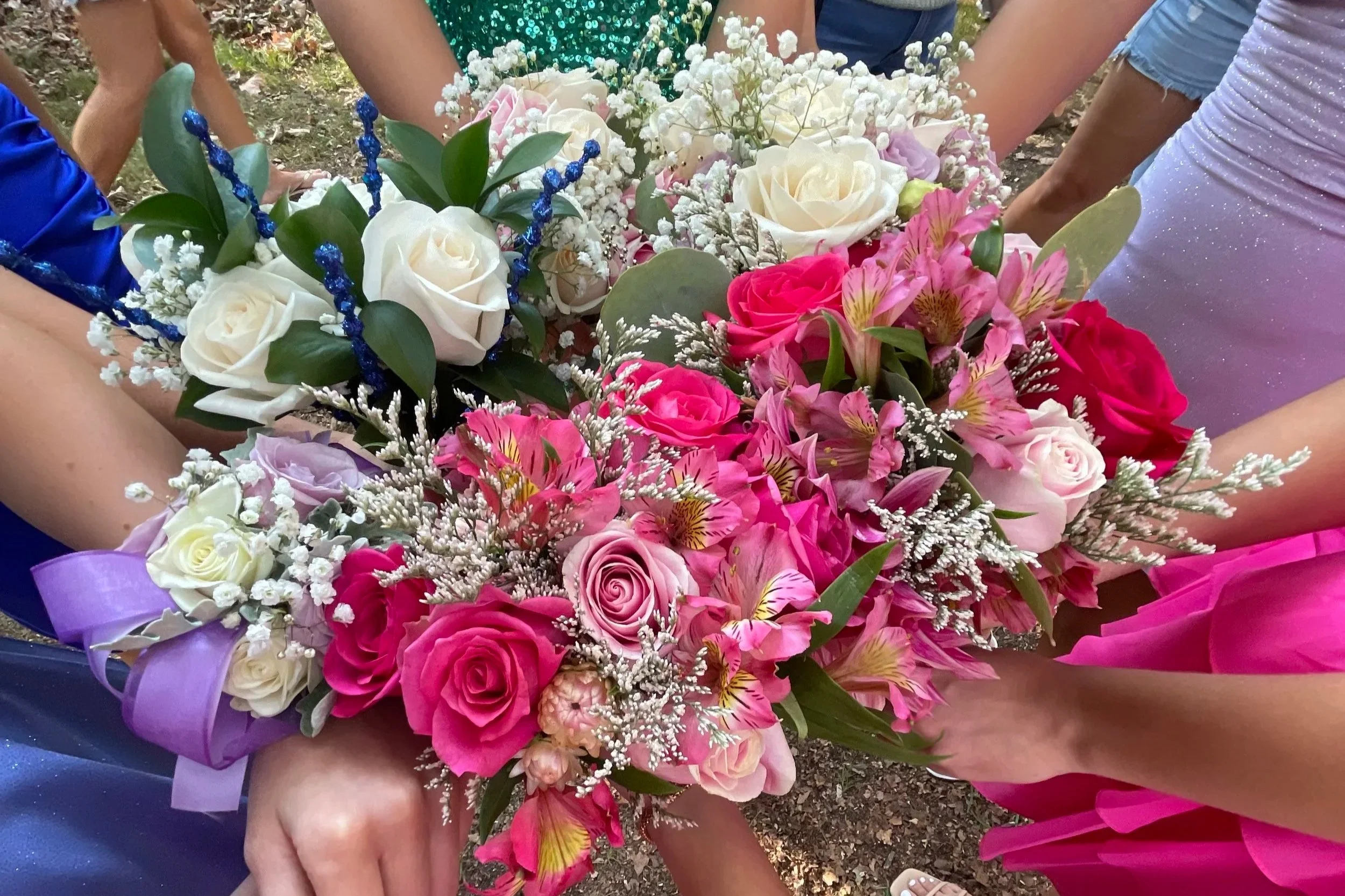 A colorful bouquet of pink, white, and purple flowers, including roses and lilies, with greenery and decorative accents, held by multiple people in celebration attire.