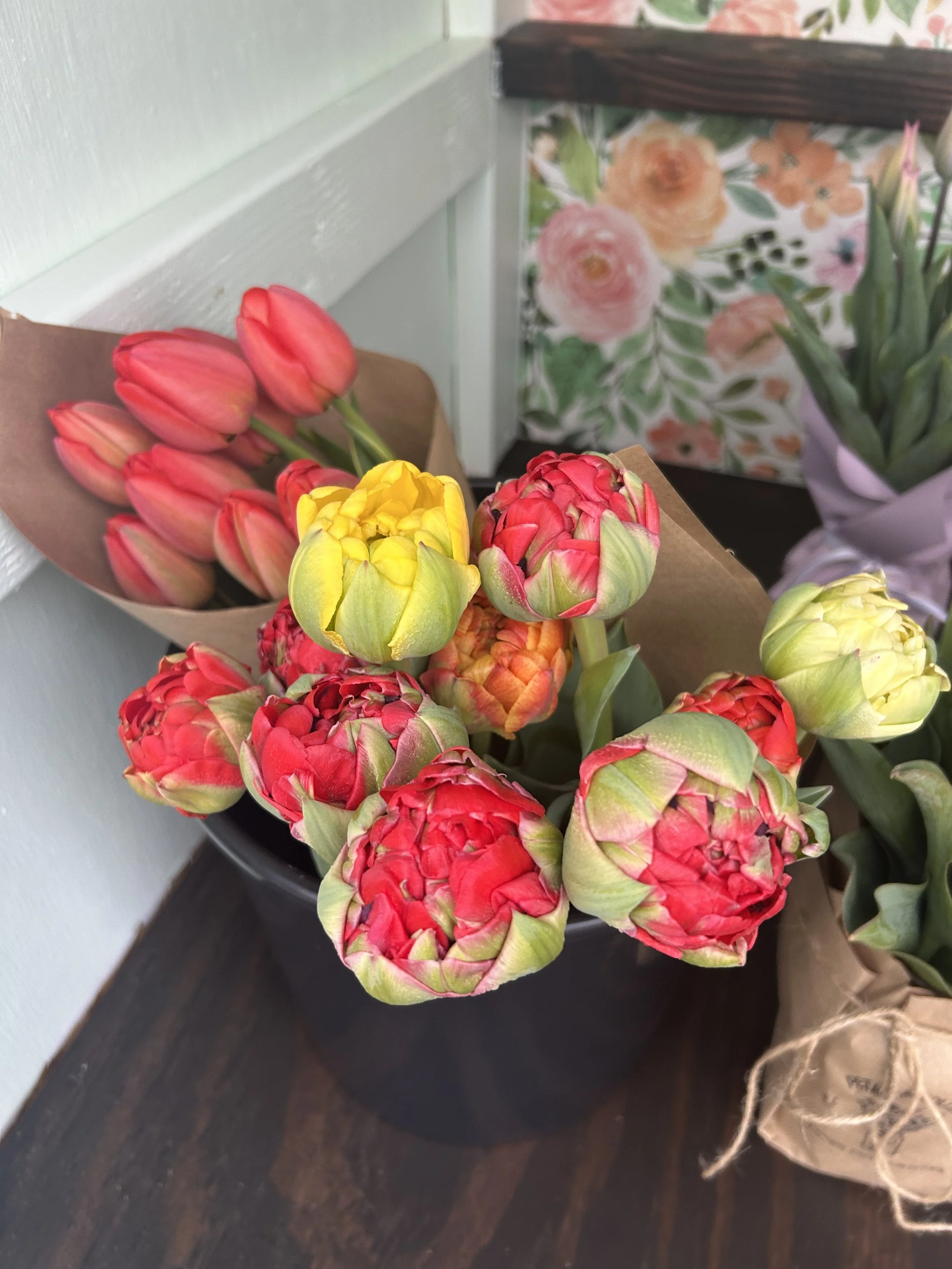 A black pot with a bouquet of colorful flowers, including pink tulips and red and yellow peonies, placed on a dark wooden surface near a light green wall.