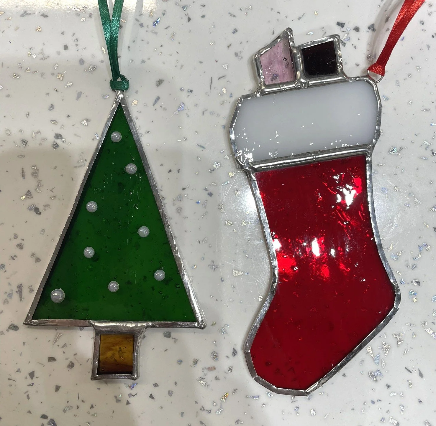 Crafted Colours' student pieces of stained glass Christmas ornaments, one shaped like a Christmas tree with green glass and decorative pearl accents, and the other shaped like a stocking with red, white, and black glass sections.