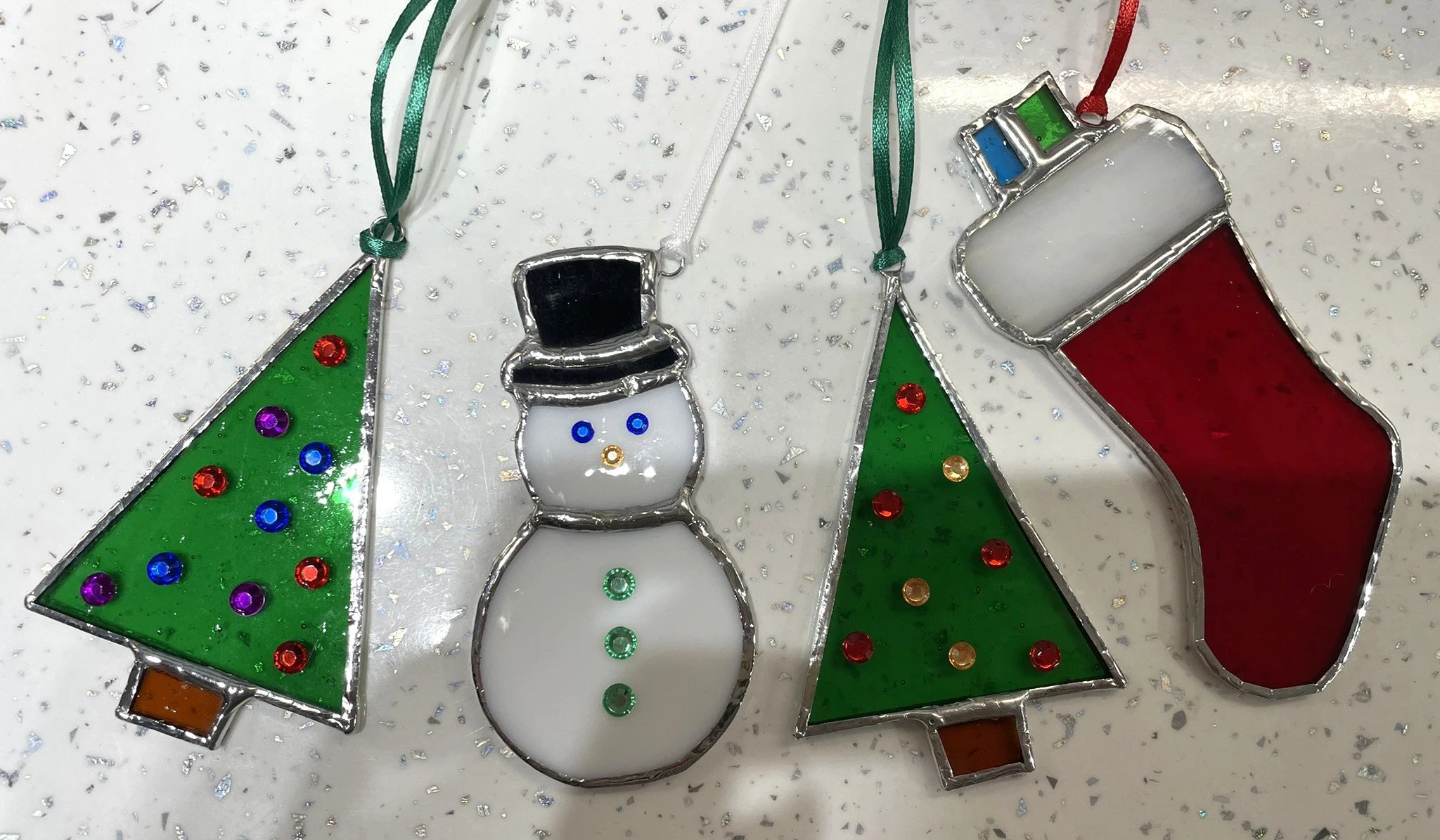 Crafted Colours' Student made stained glass Christmas ornaments: two Christmas trees with colorful decorations and a snowman with a black top hat, all hanging on ribbons.
