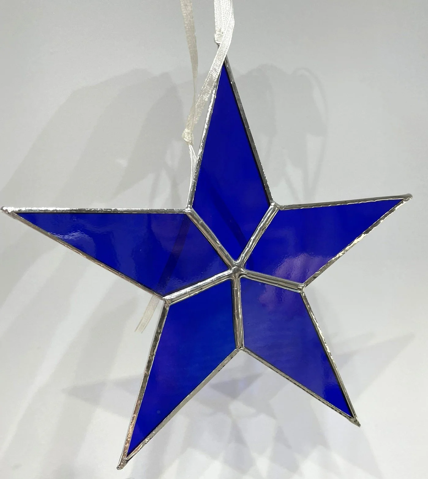 Crafted Colours' student piece of a decorative blue stained glass star ornament hanging by a white ribbon.