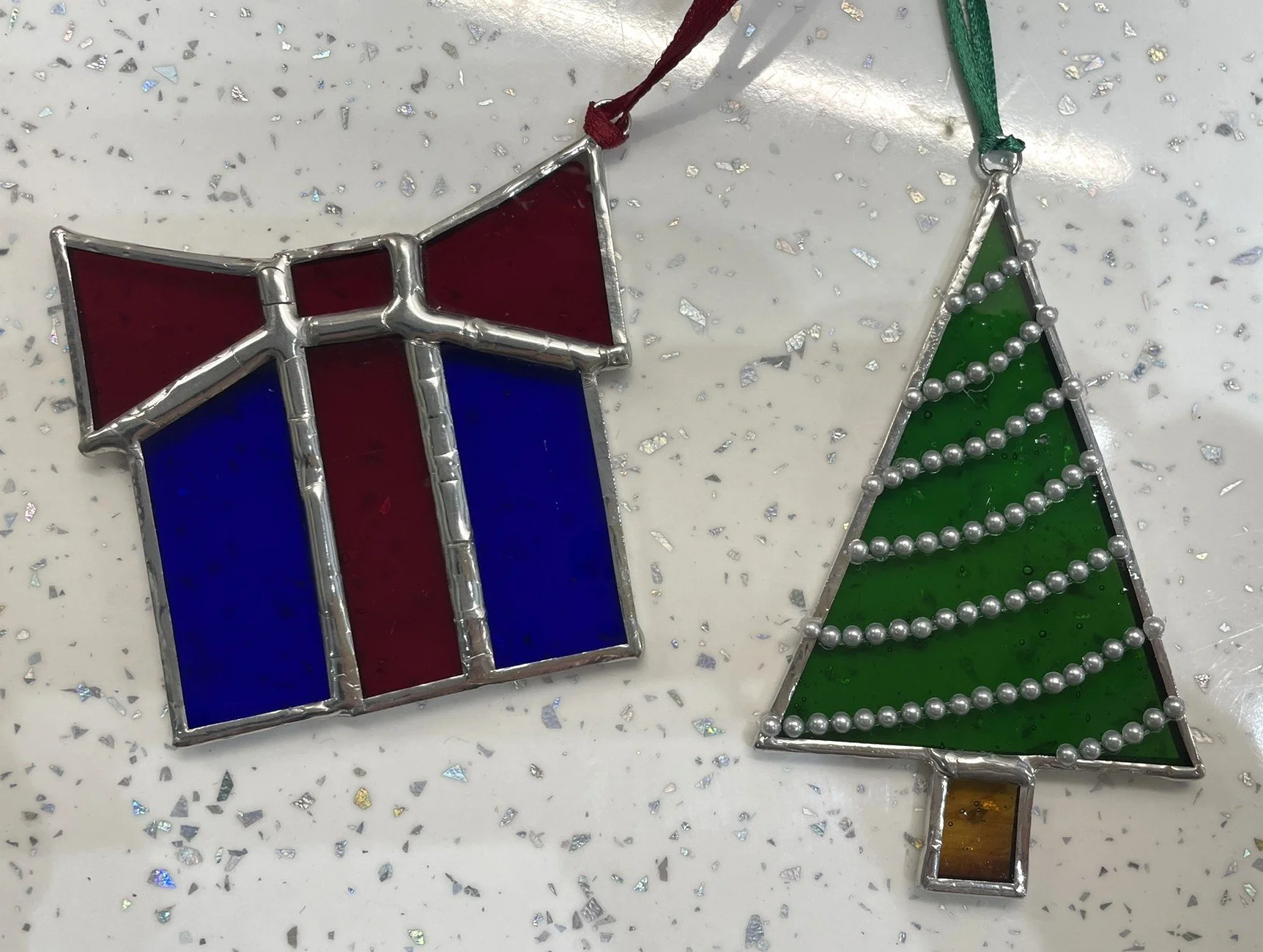 Crafted Colours' student pieces comprising two stained glass Christmas ornaments: one in the shape of a wrapped present with red and blue glass, and the other in the shape of a Christmas tree with green glass and silver bead garlands.