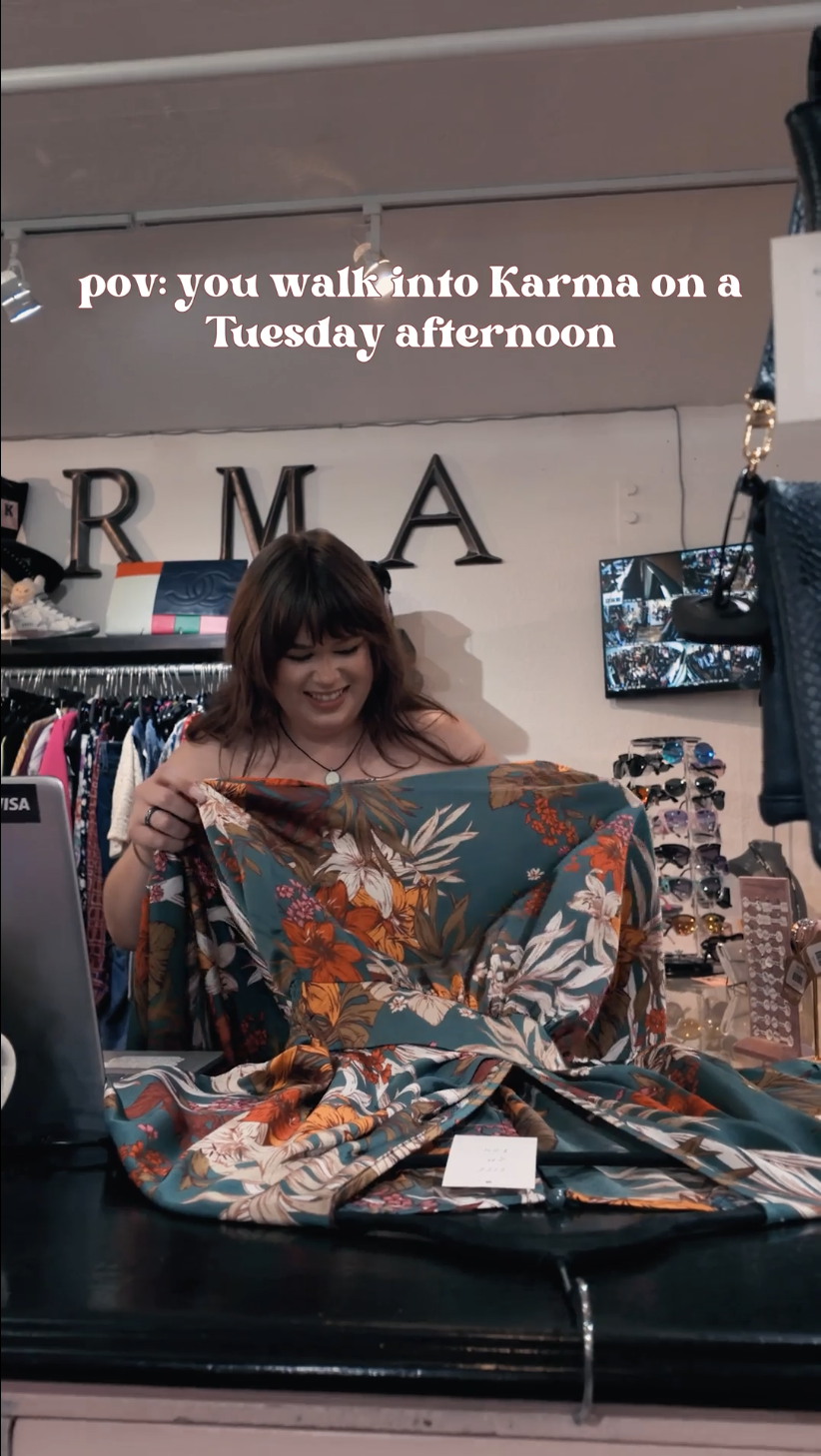 A woman with shoulder-length brown hair smiling as she looks at a colorful patterned fabric in a store. Behind her are sunglasses on display and decorative letters on the wall spelling 'KARMA'.