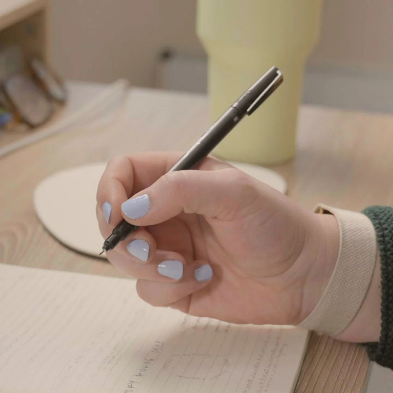 Person with light nail polish writing in a notebook with a pen on a wooden desk, with a green cup and some blurred items in the background.