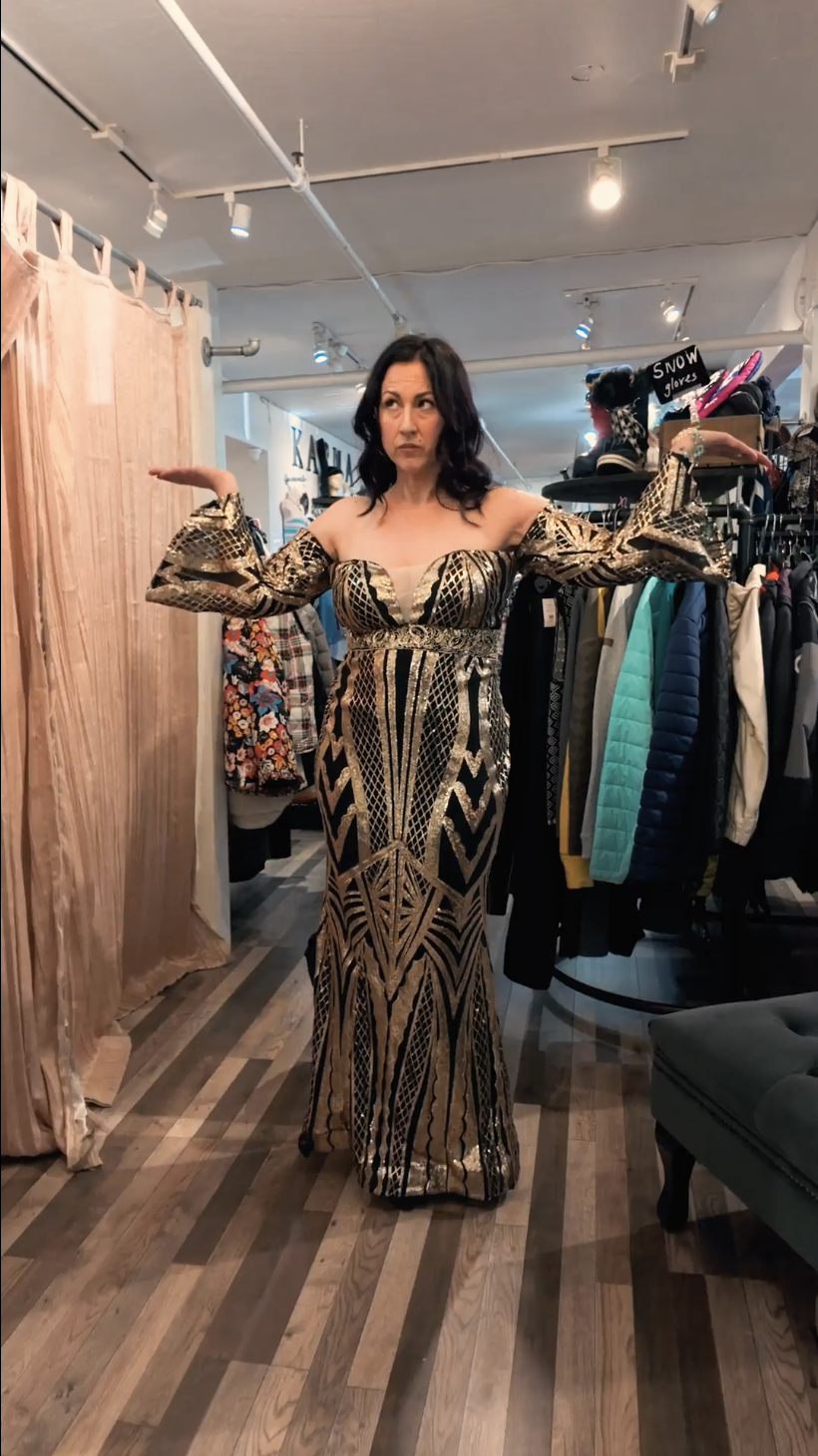 Woman trying on a long, patterned metallic dress in a clothing store, with colorful coats and accessories on racks behind her.