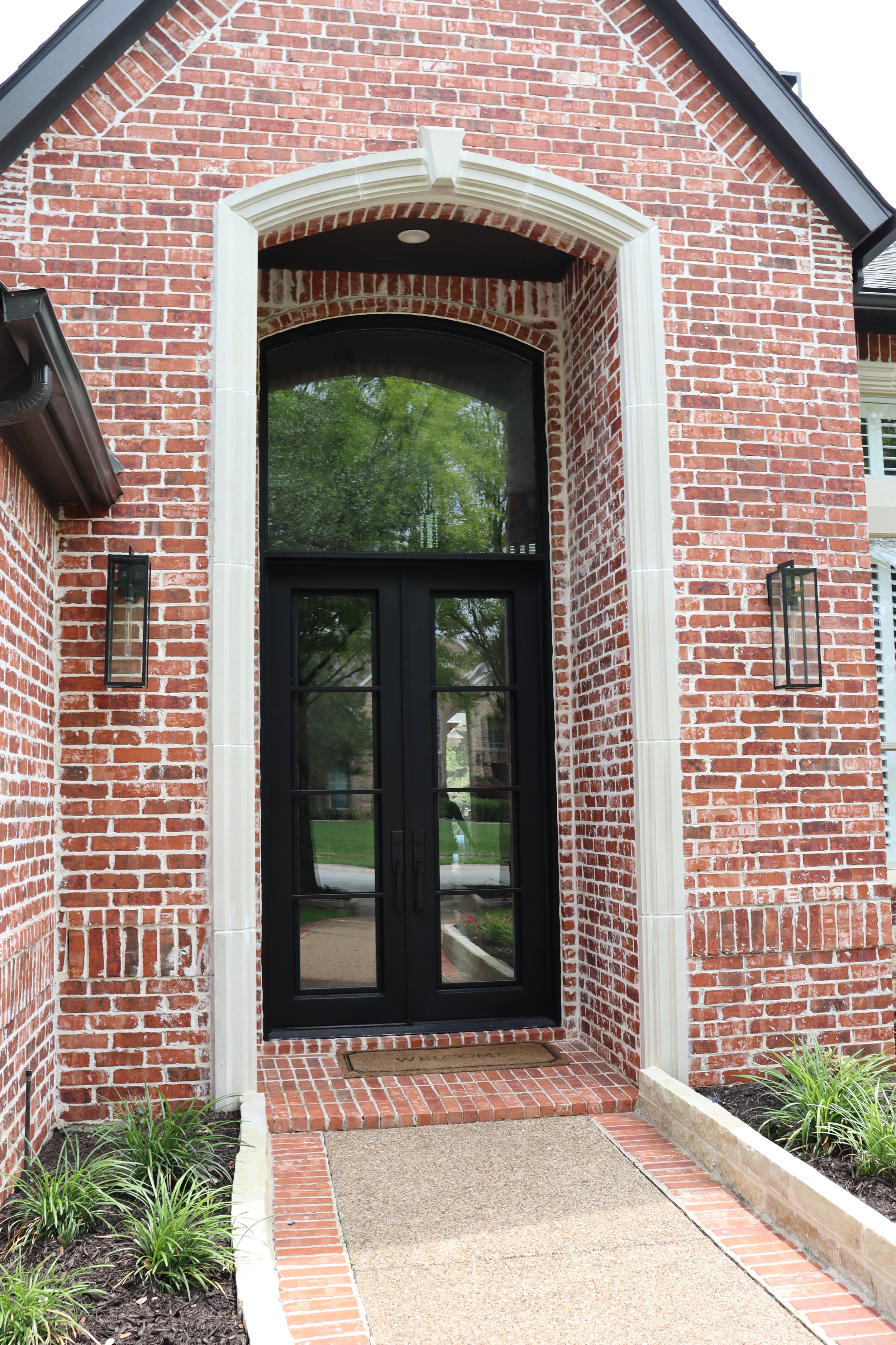 The front door of a house with brick exterior, black double doors with glass panes, an arched window above, and decorative white trim. A doormat is placed in front, and garden beds with plants line the walkway.