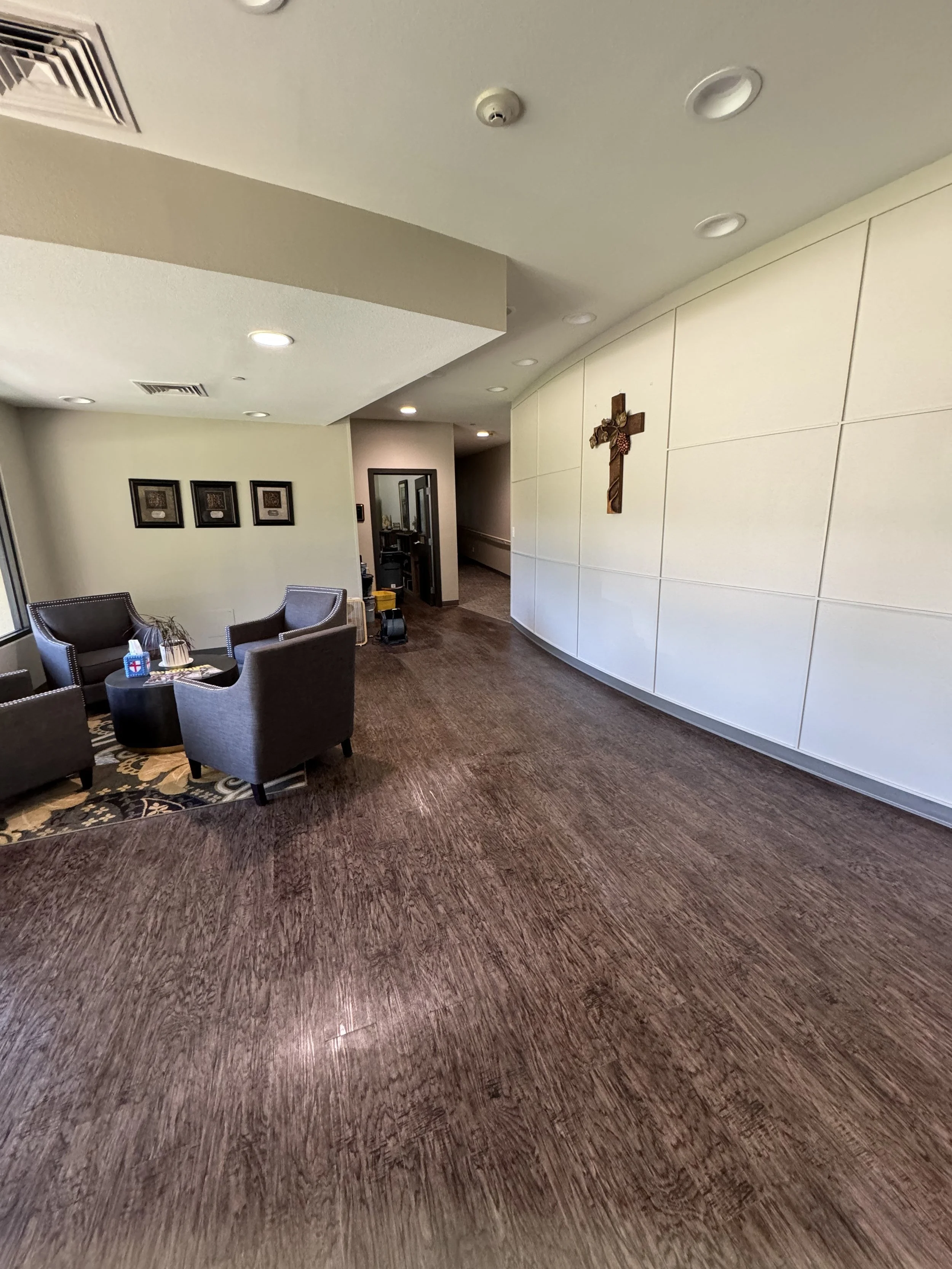 Interior of a room with beige walls, framed artwork, a cross on the wall, chairs around a small table, and a hallway in the background.