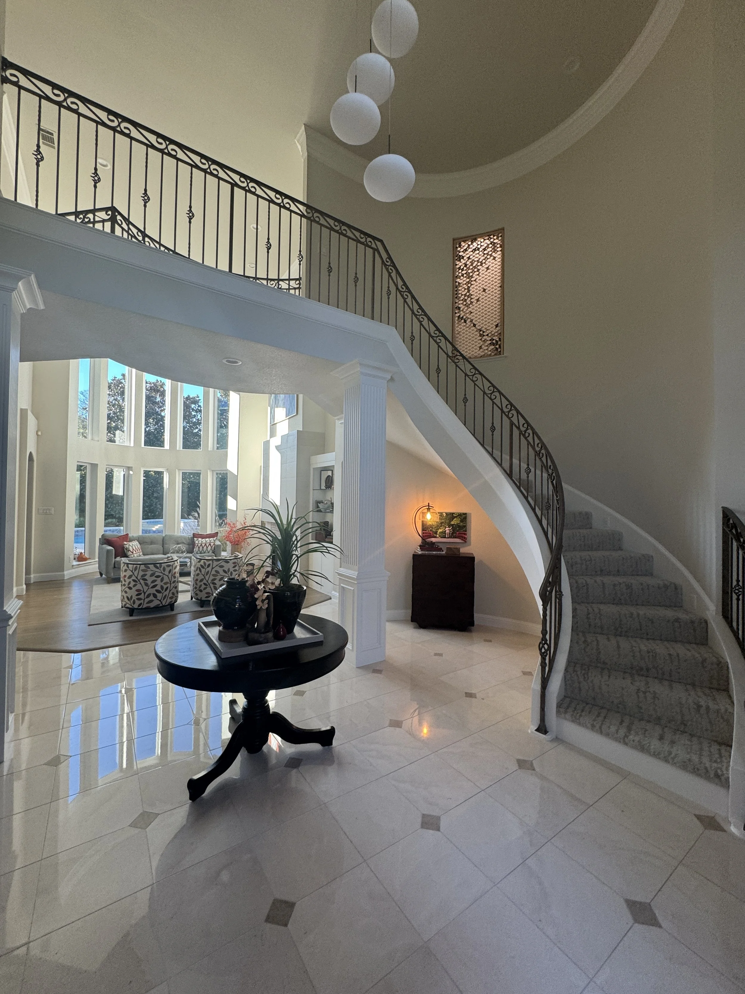 Interior of a large home with a curved staircase, black wrought iron railing, and a chandelier with five spherical white lights hanging from the ceiling. The floor is tiled, and there is a round black table with decorative vases. The living area has large windows, a sofa with patterned cushions, and a small table with a lamp near the stairs.