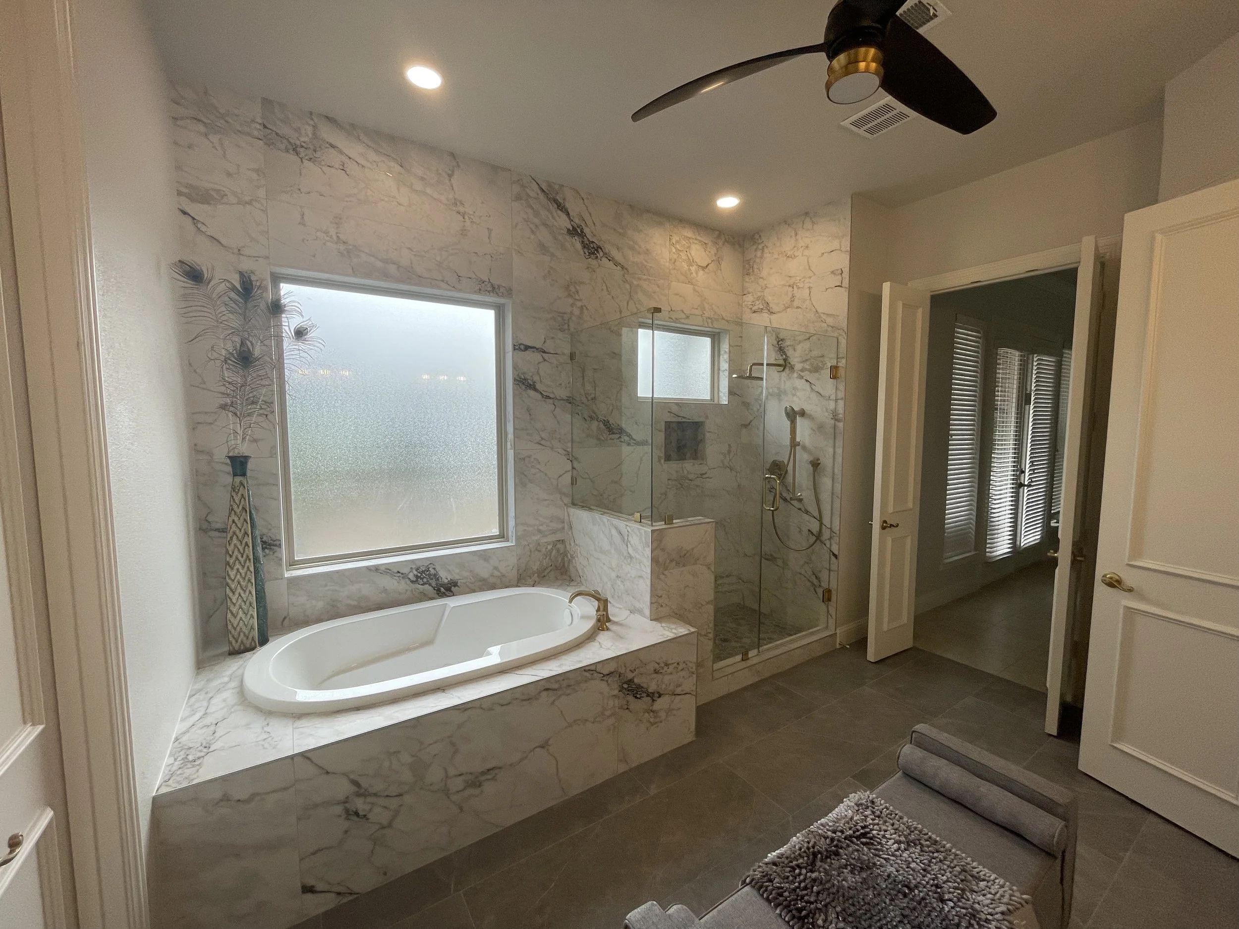 A bathroom with a corner bathtub, marble tiles, and a glass-enclosed shower. There is a frosted window, a decorative vase with feathers, and a door leading to a room with window blinds.