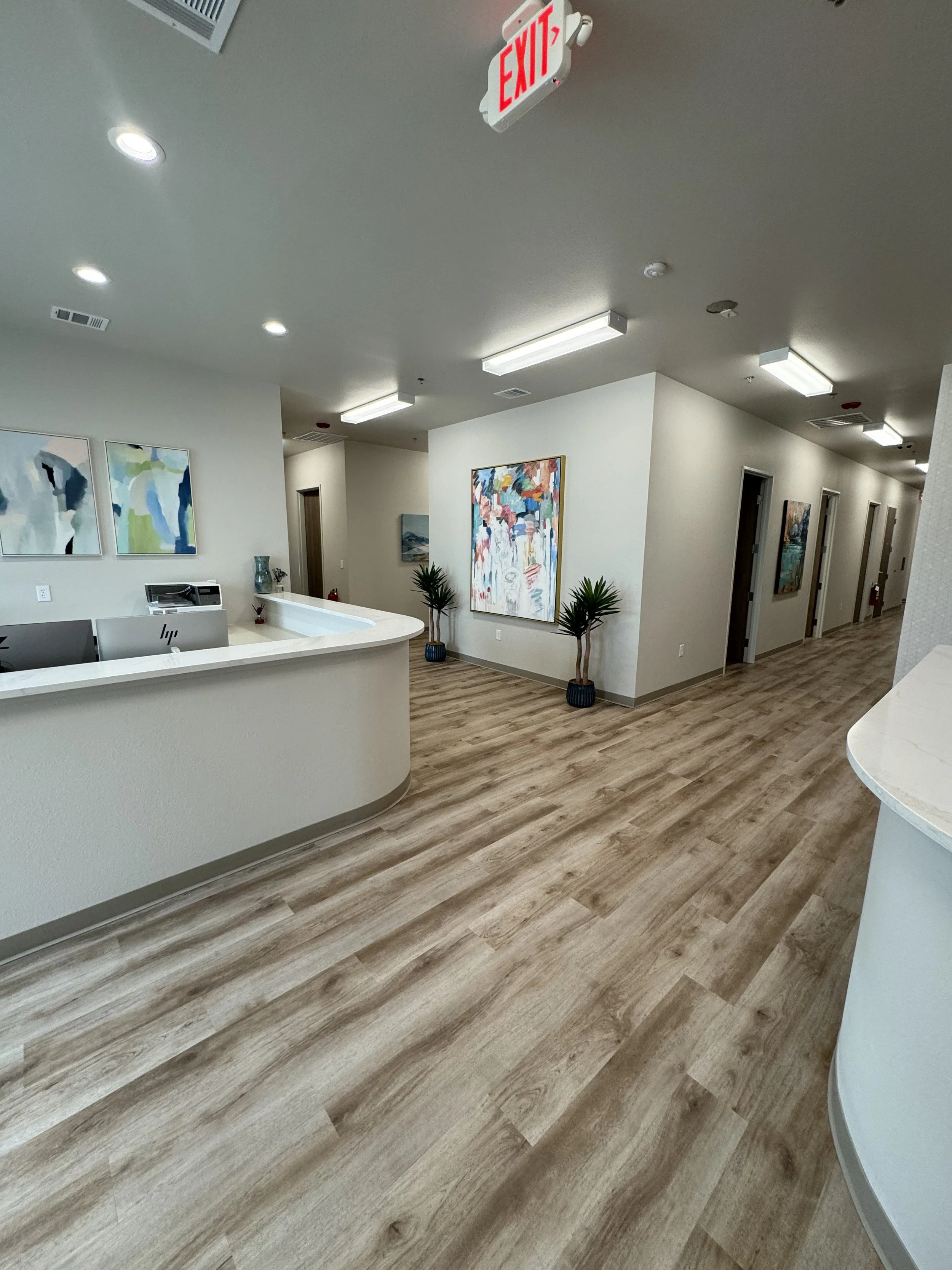 Interior view of an office or medical facility hallway with wooden flooring, white walls, and multiple art pieces hanging. There are some potted plants near the wall, ceiling lights, and an exit sign hanging from the ceiling.