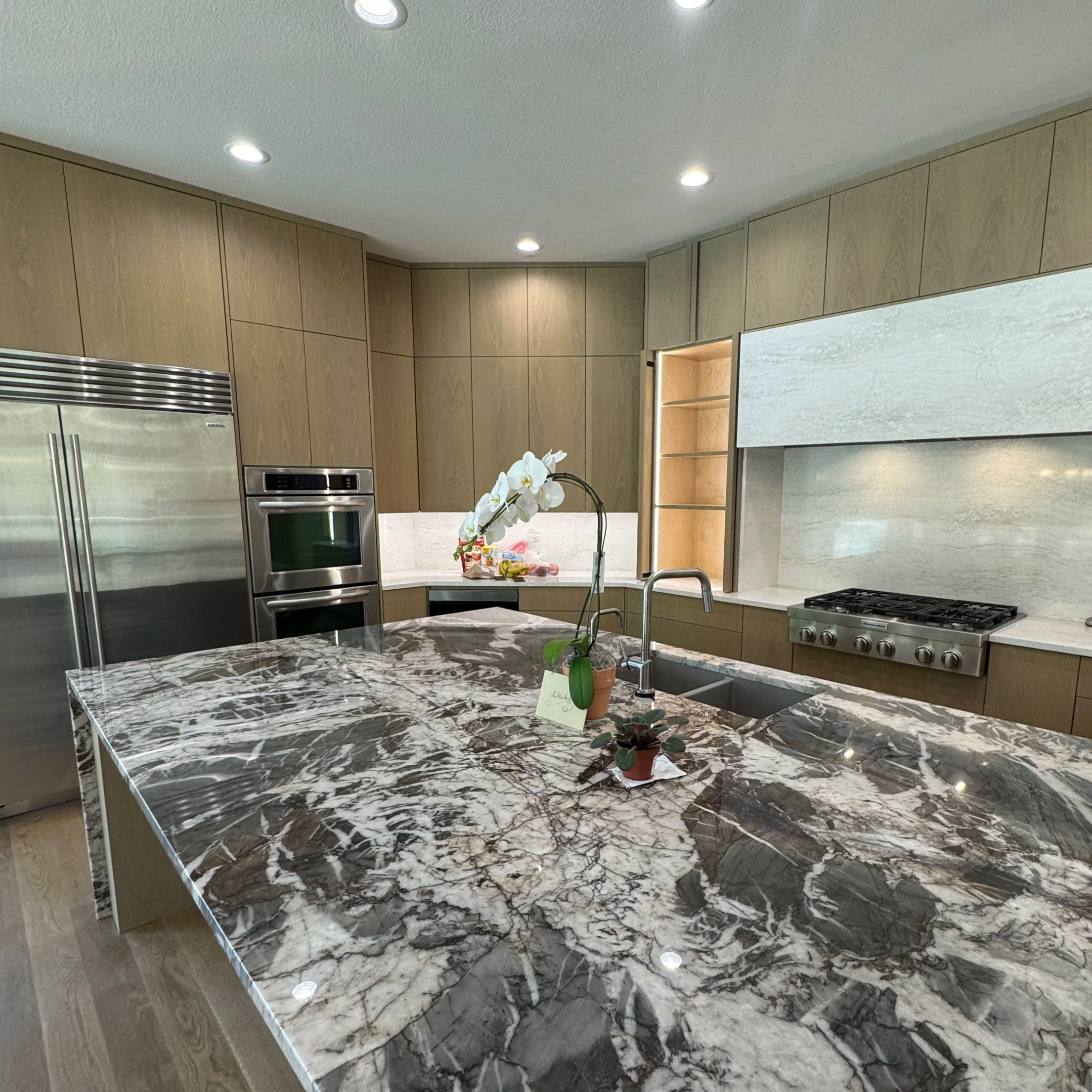 A kitchen with a black and white marble countertop, wooden cabinets, stainless steel refrigerator and oven, a flower arrangement on the counter, and bright lighting from ceiling fixtures.