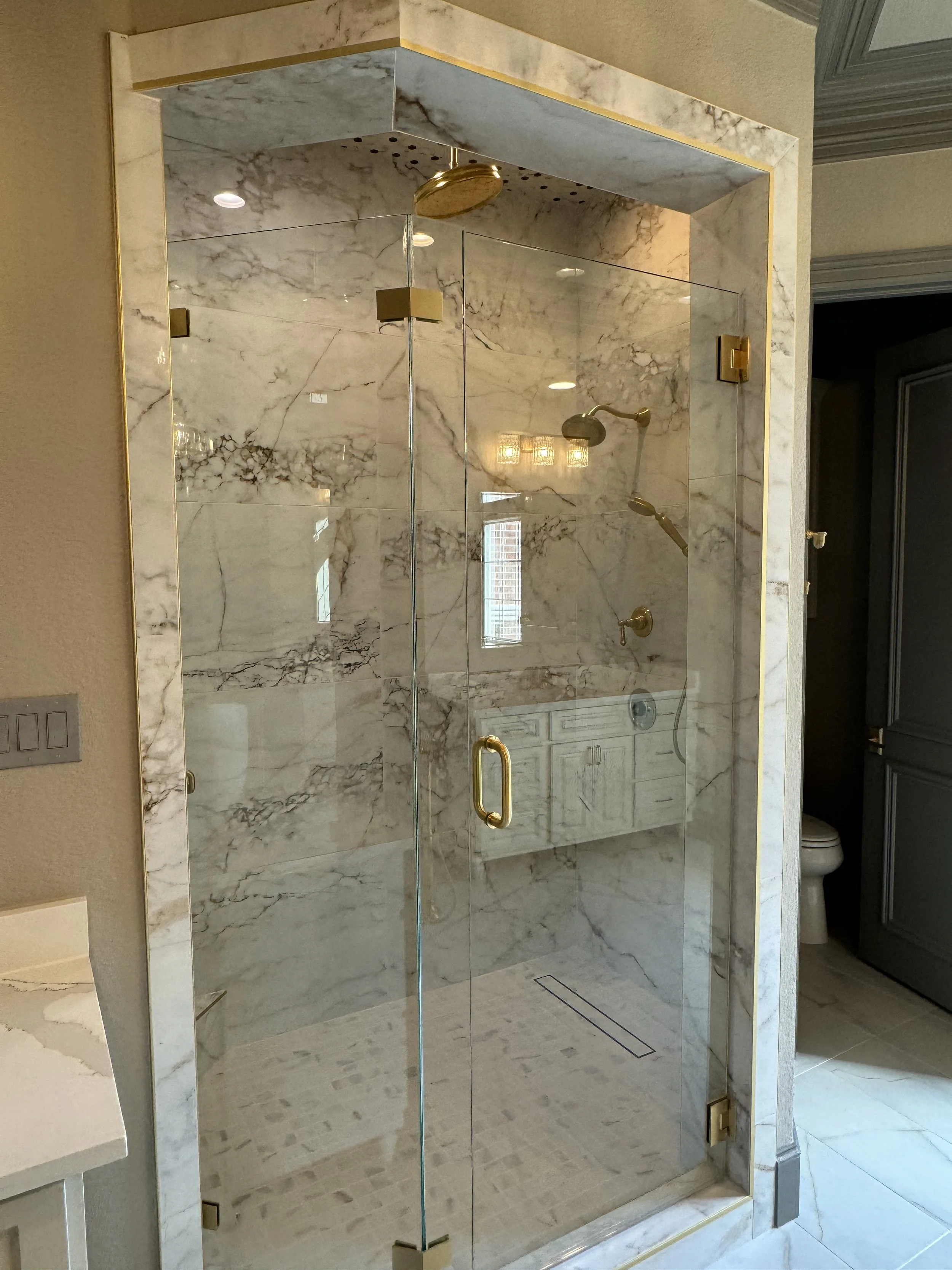 A modern enclosed shower with marble walls and floor, equipped with gold fixtures and a glass door, located in a bathroom.