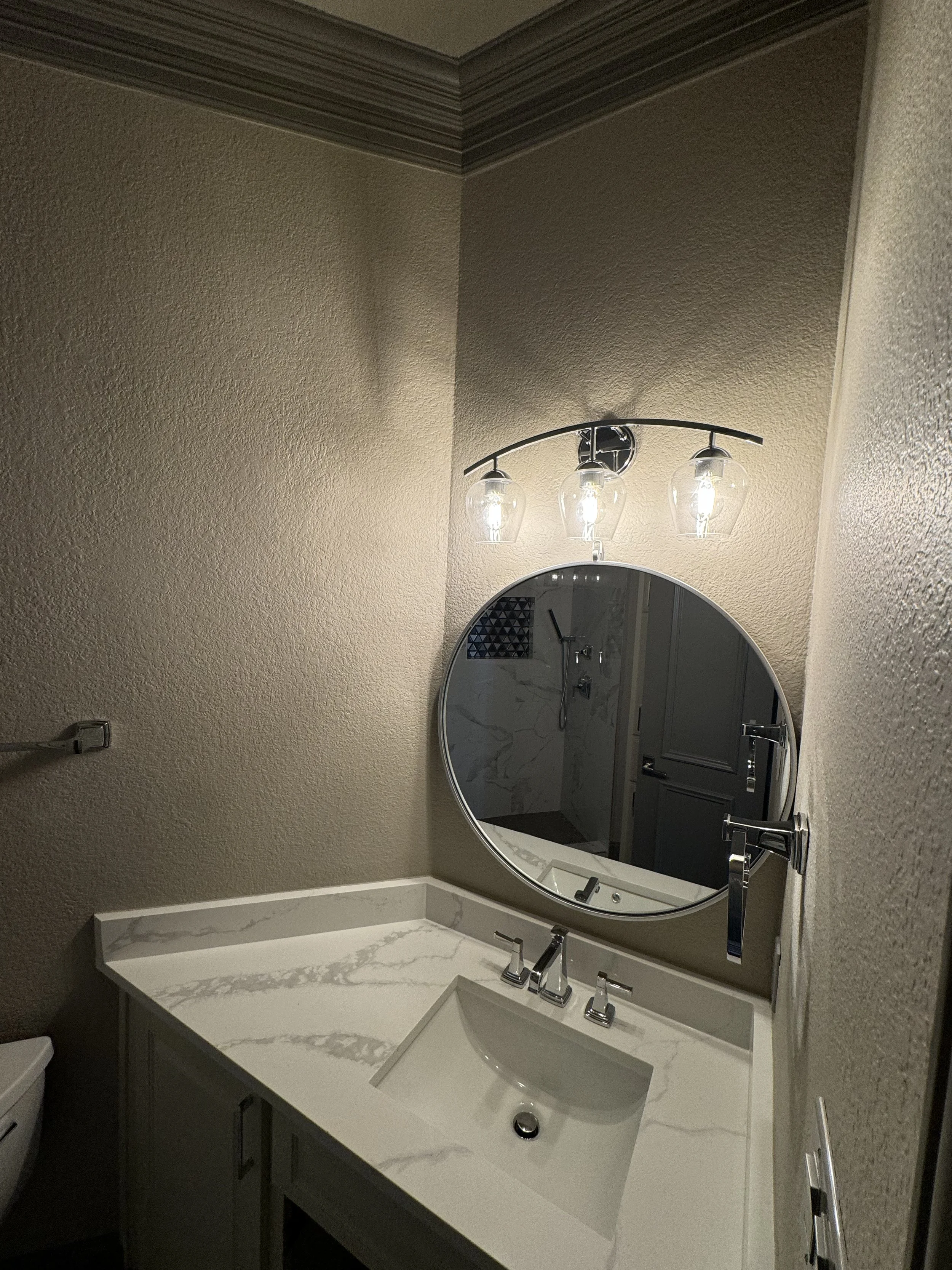 Bathroom vanity with a white marble countertop, an oval mirror, a three-light vanity fixture, and a textured beige wall.