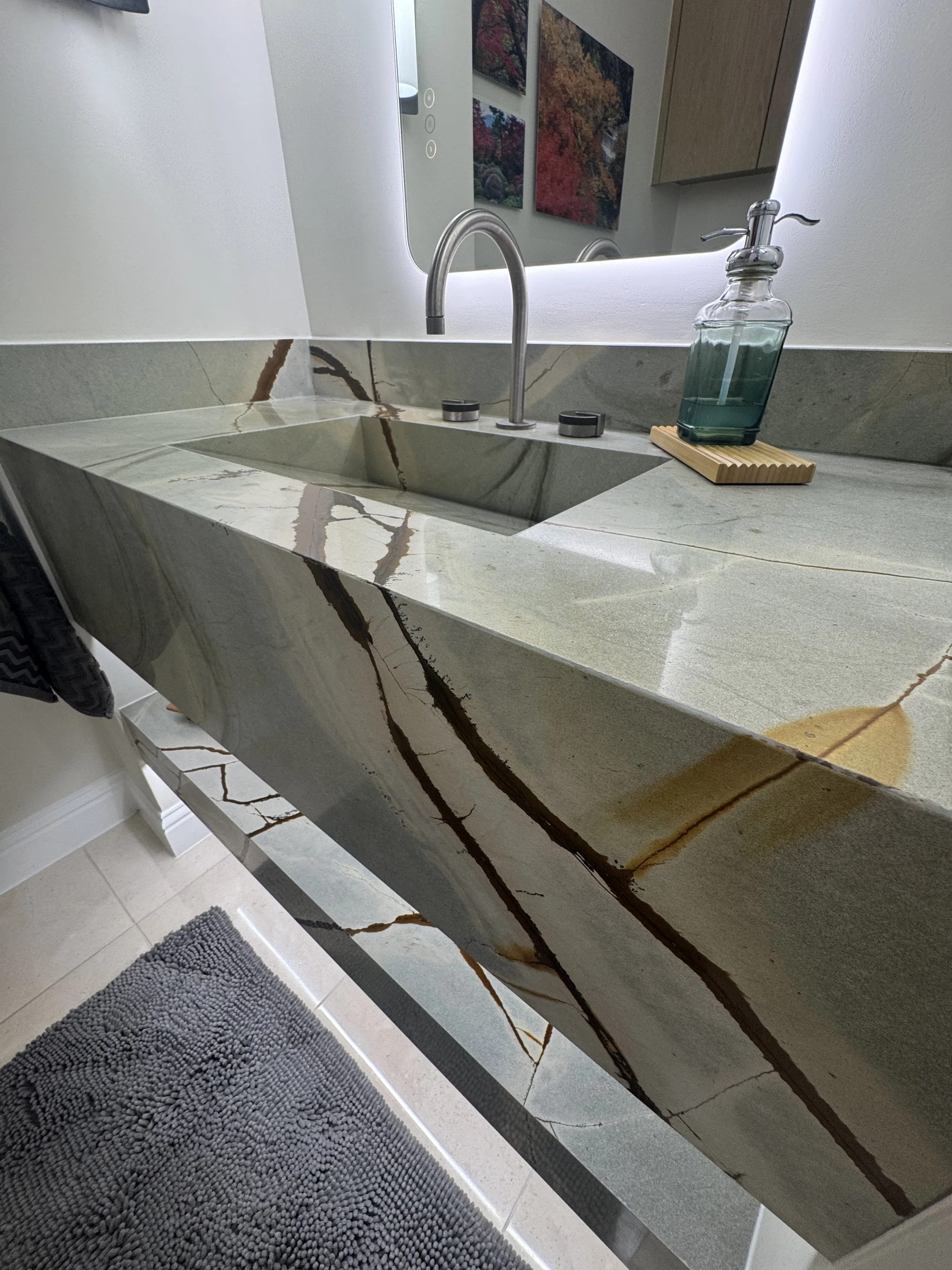 A bathroom sink with a marble vanity top, a silver faucet, a bottle of green soap with a wand, and a mirror above the sink reflecting some colorful artwork on the wall.