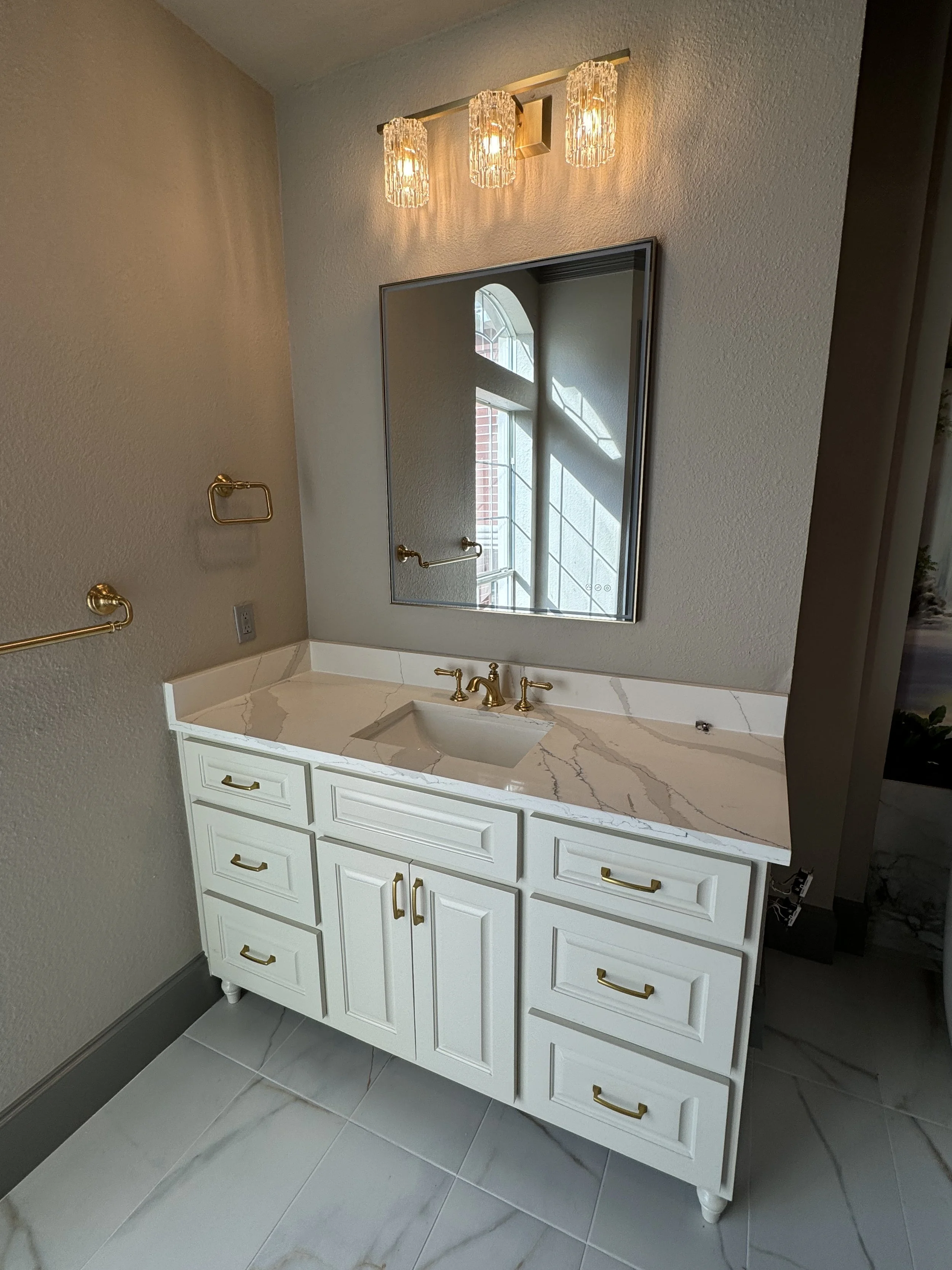 A white bathroom vanity with gold handles, marble countertop, gold faucet, a mirror above, three pendant lights, gold towel rings, and gray tile flooring.