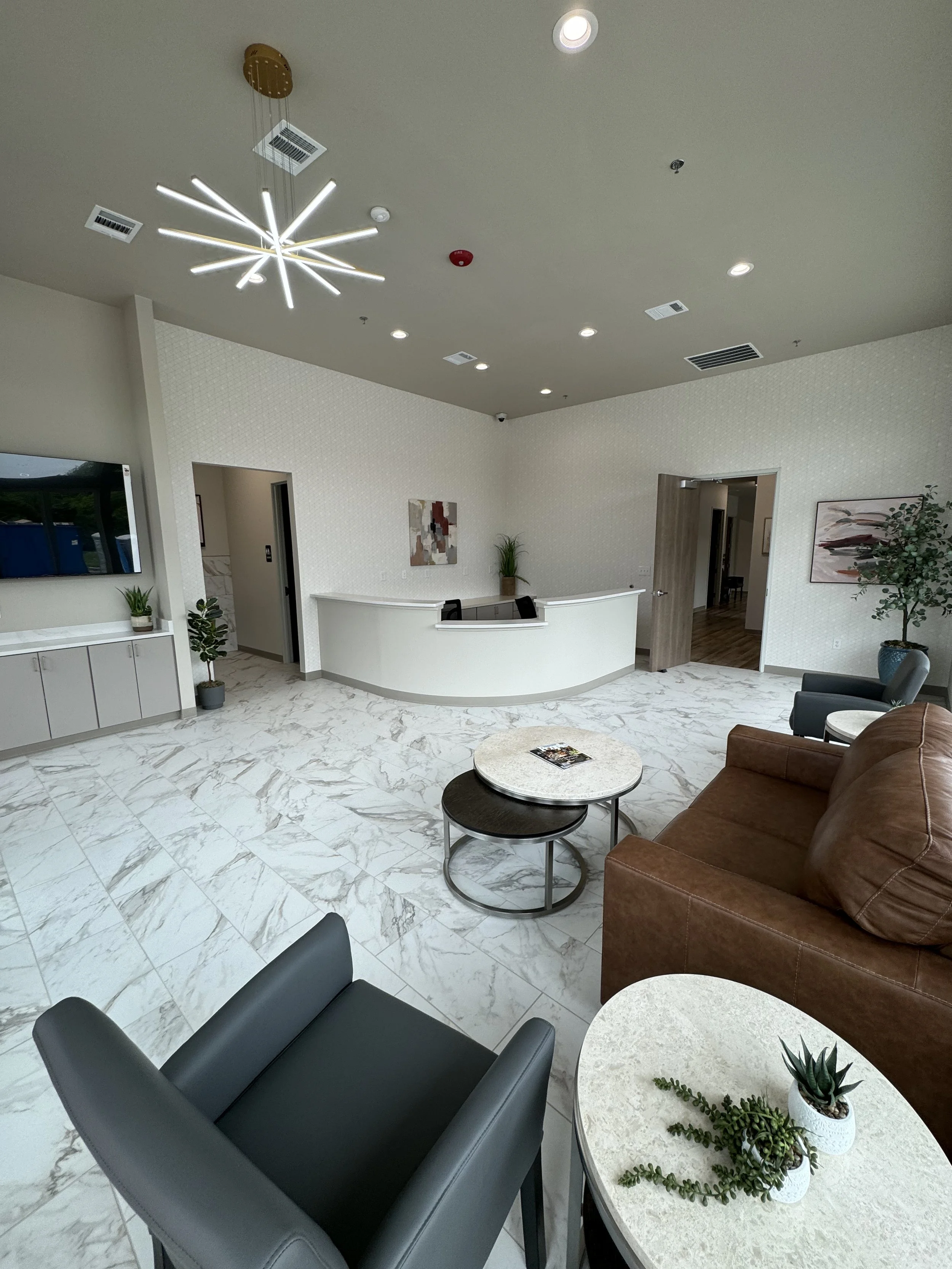 A modern waiting area or lobby with white marble flooring, a curved reception desk, brown leather sofa, black chairs, round tables with potted plants, and abstract artwork on the wall. Ceiling features a unique modern light fixture and recessed lighting.