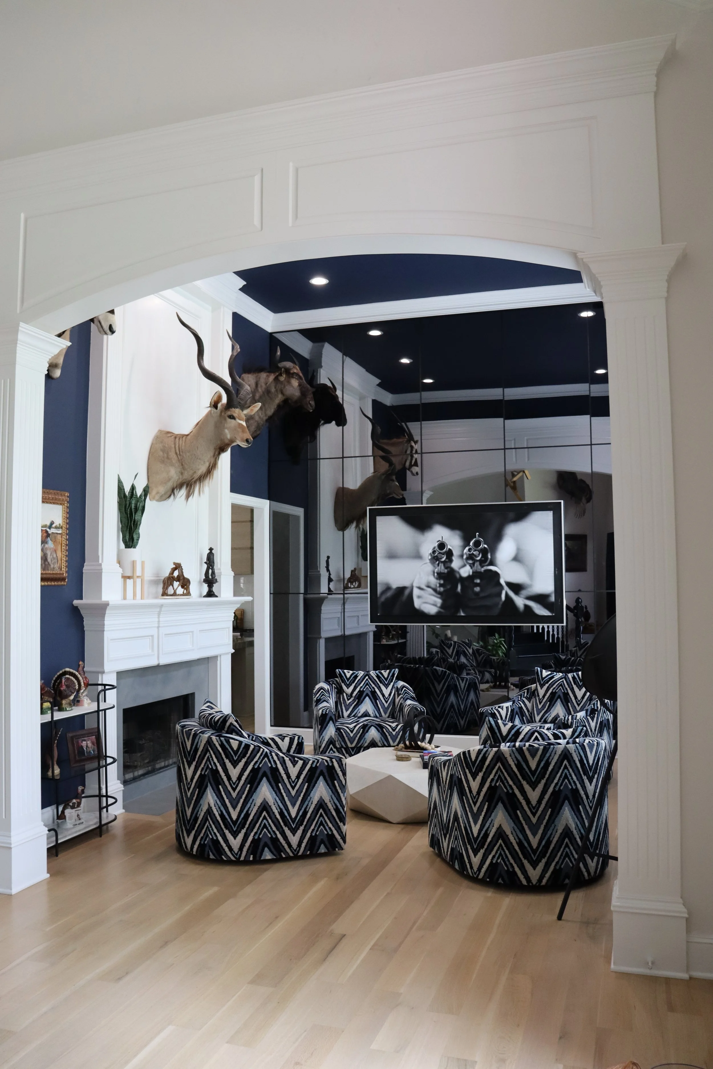 Living room with a fireplace, mounted animal heads, patterned chairs, a wall-mounted television, and various decorative items.