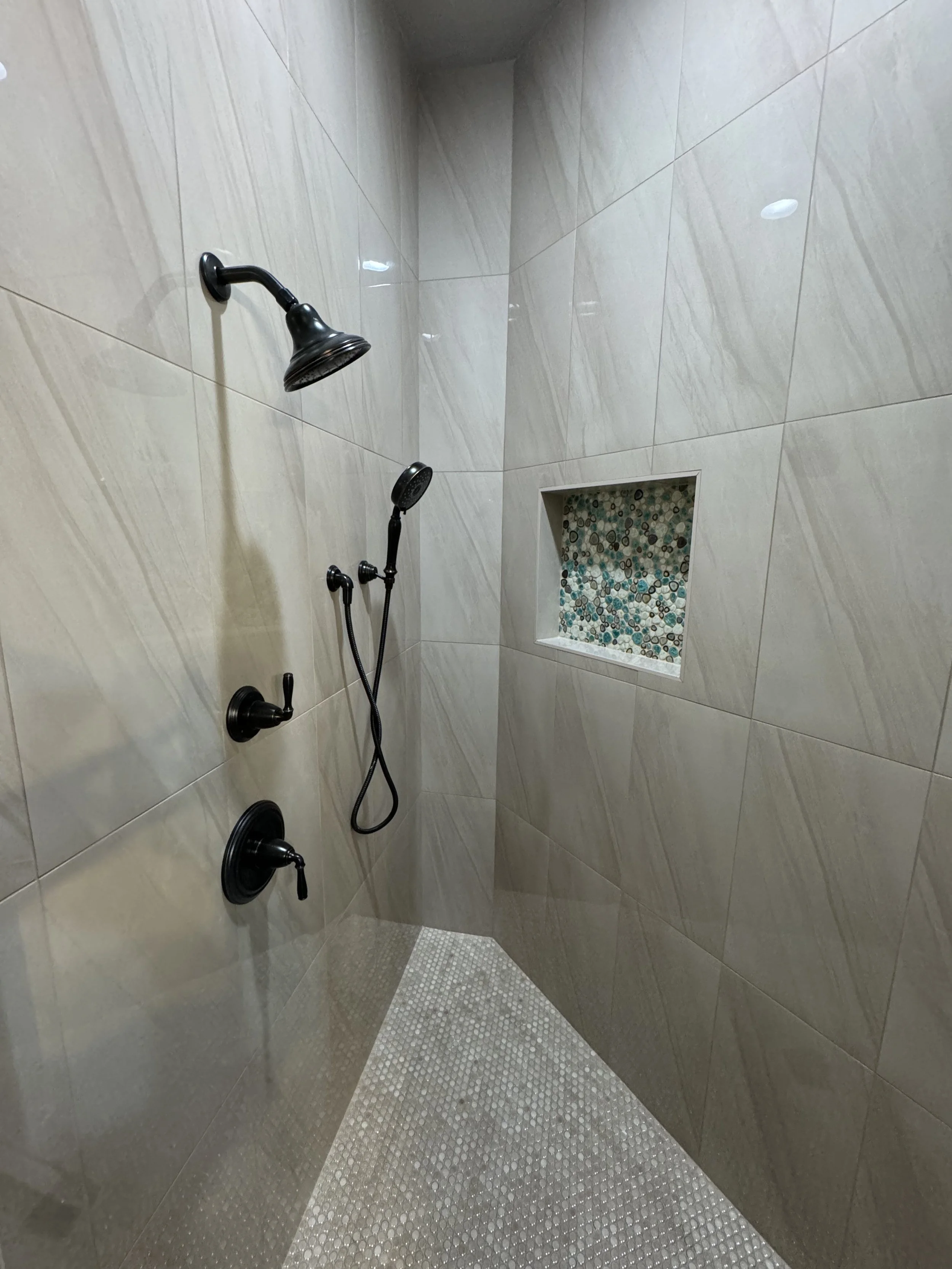 A modern walk-in shower with beige tiled walls, a black showerhead, a handheld shower wand, a built-in niche with decorative pebbles, and a textured non-slip shower floor.