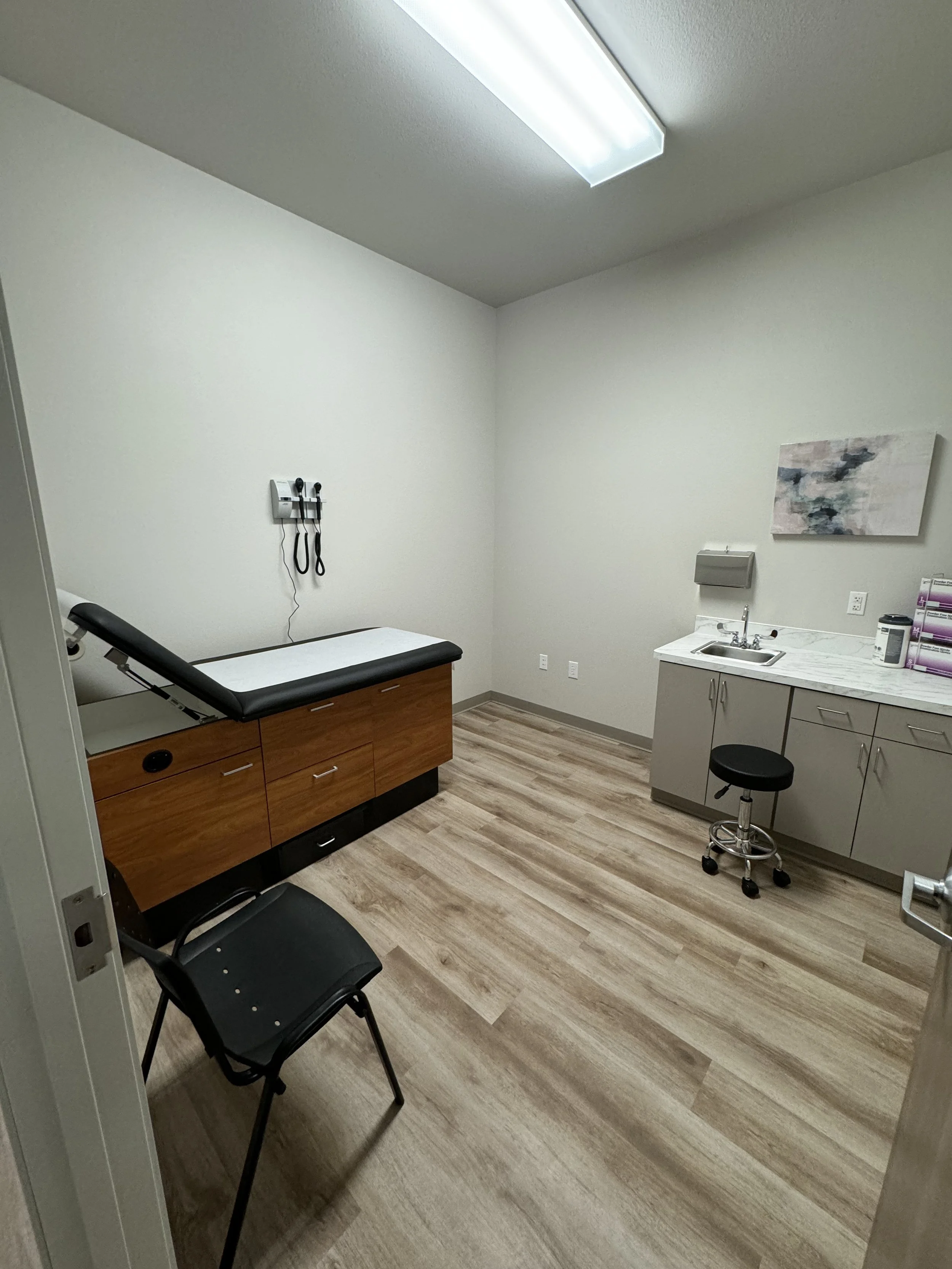 A medical examination room with an examination table, wall-mounted medical devices, a sink with cabinets, a piece of abstract art, and chairs.