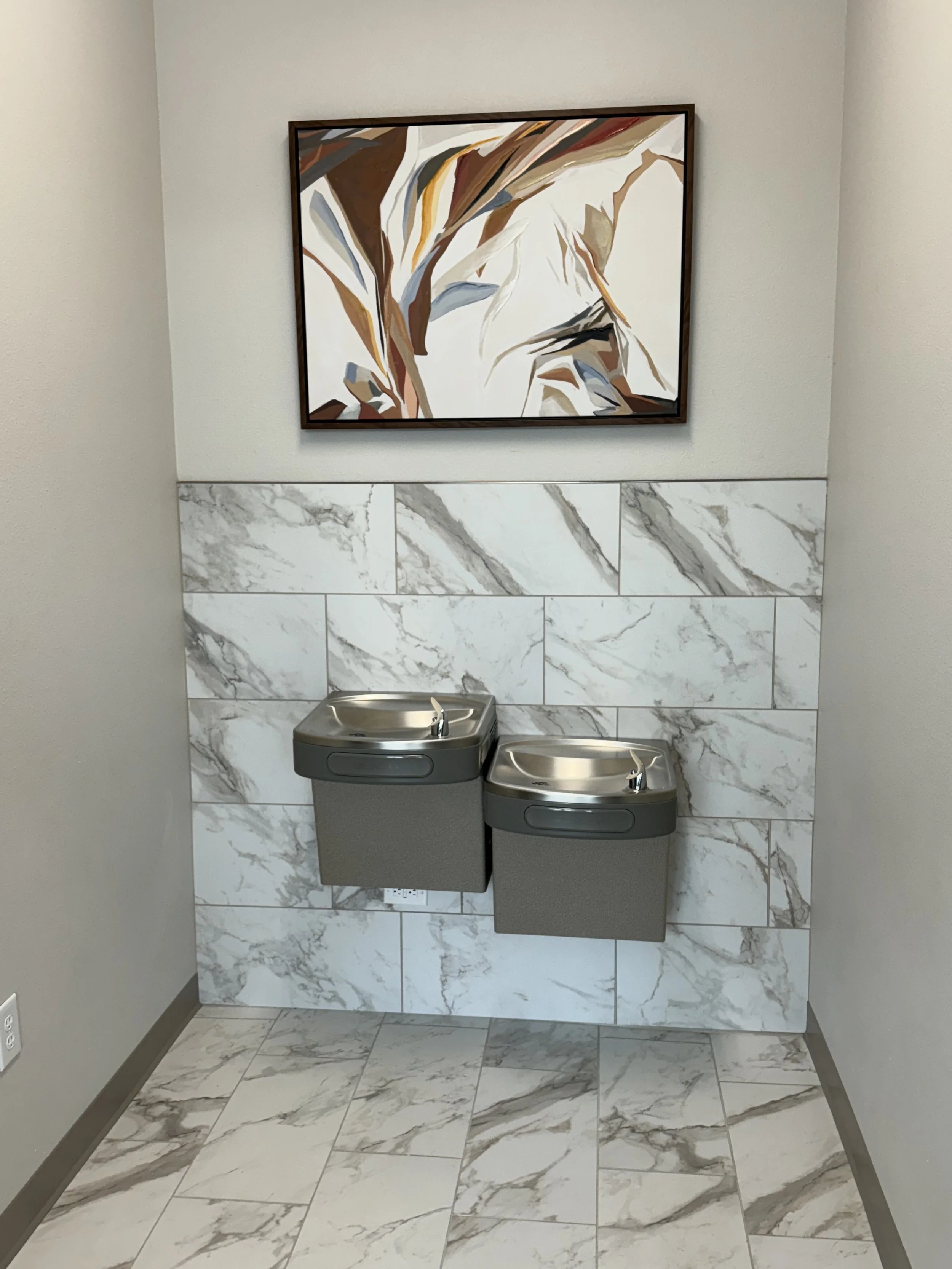 A bathroom with marble tiles on the wall and floor. Two gray hand sanitizer dispensers are mounted on the wall under a framed abstract art piece with earth tones.