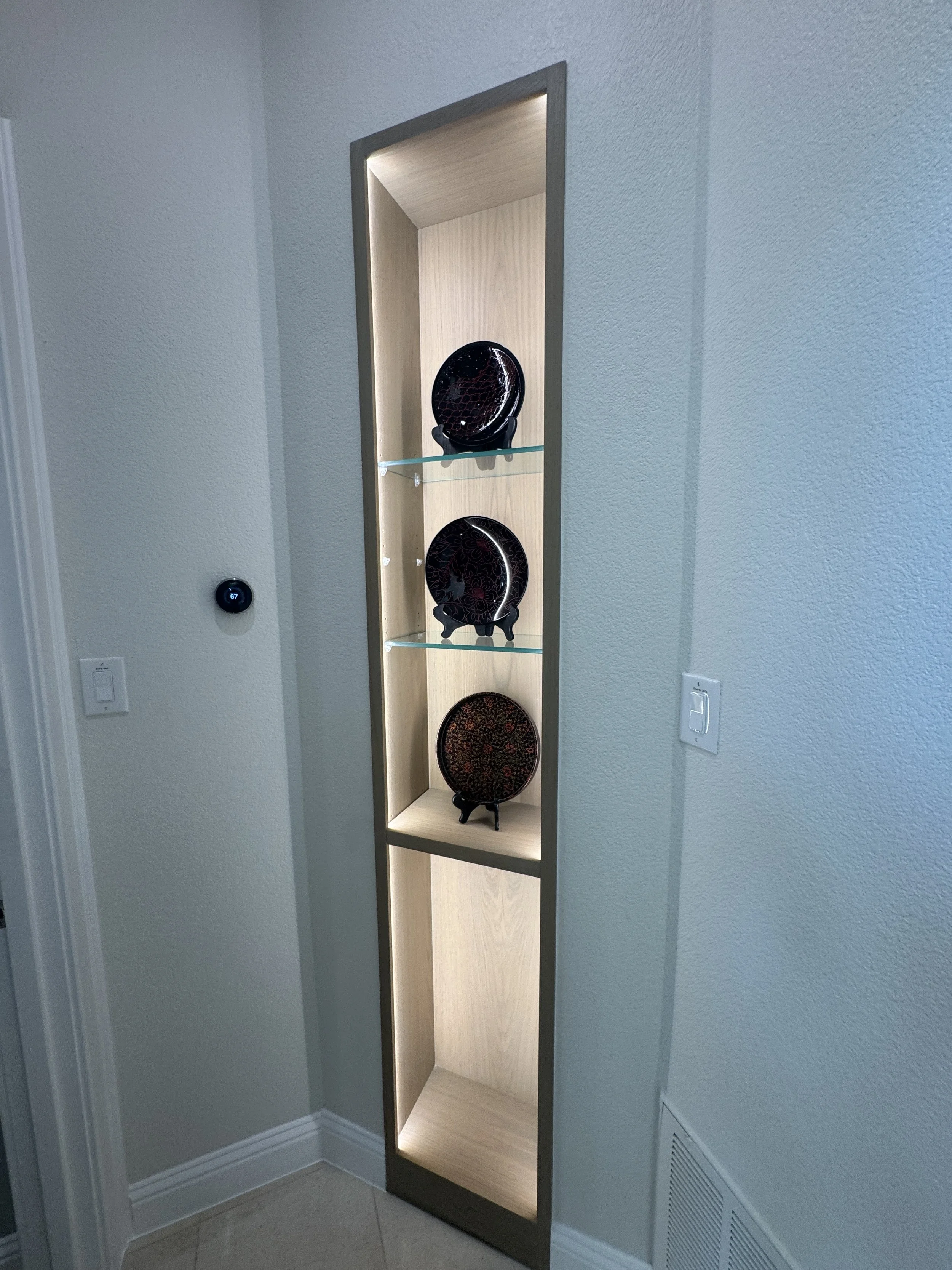 Display cabinet with three shelves containing decorative plates, situated in a corner of a room with white walls and a wood-colored interior