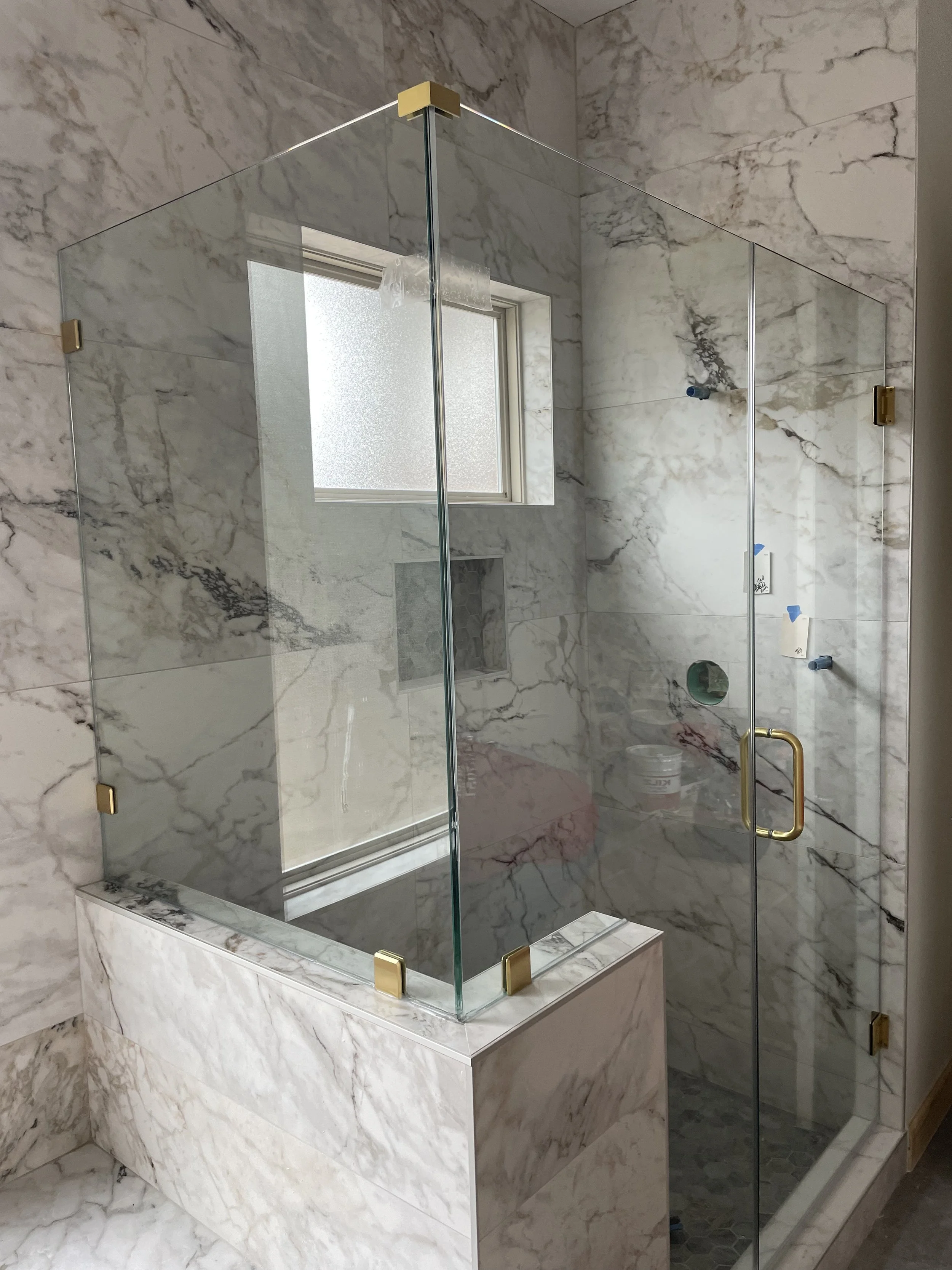 A marble-walled bathroom shower with a glass enclosure and brass hardware, featuring a frosted window and a showerhead pipe connection.